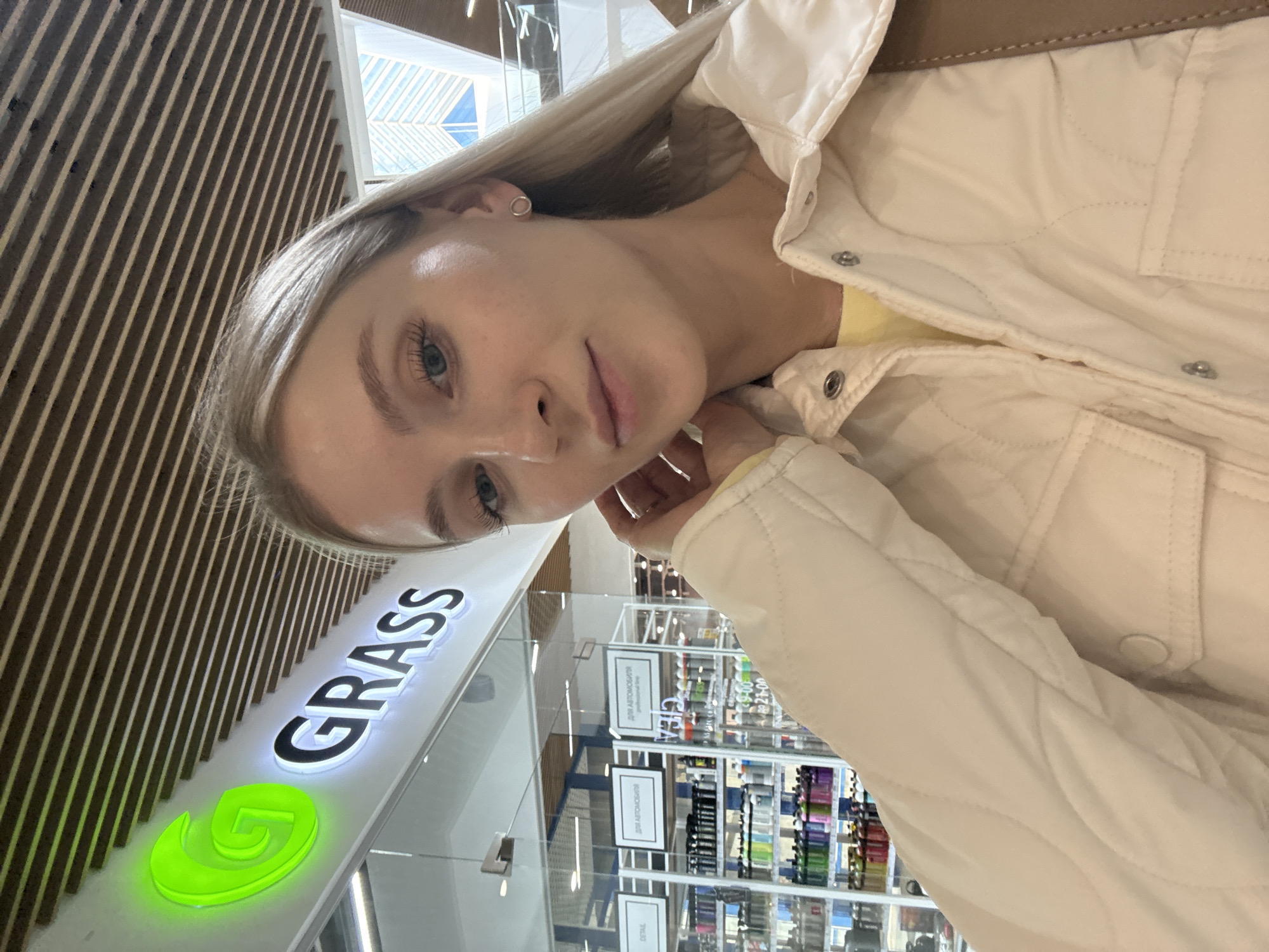 A young woman with blonde hair is taking a selfie in front of a GRASS store. The store appears to be located in a shopping mall in Izhevsk, Russia. The woman is wearing a cream-colored jacket and is looking directly at the camera. Behind her, shelves are stocked with various products. The GRASS logo is illuminated with green neon. The background includes a wall with a striped texture.