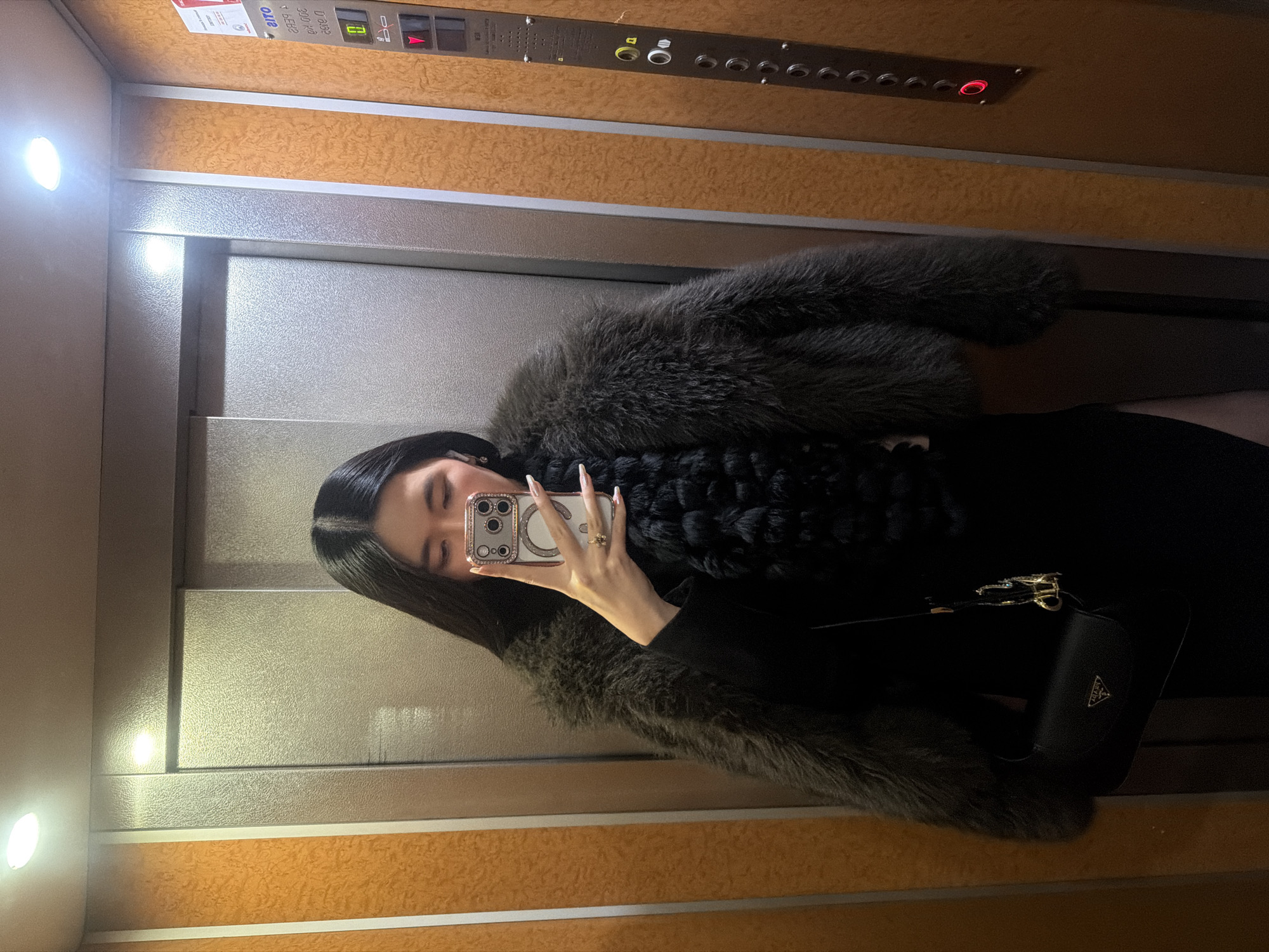 A woman in an elevator takes a selfie with a smartphone. The woman is wearing a fur coat and a black dress, and she is carrying a black Prada handbag. The elevator has a control panel with buttons and indicator lights. The background is textured with shades of brown and gray. No specific location in Paris, France, can be determined from the image.