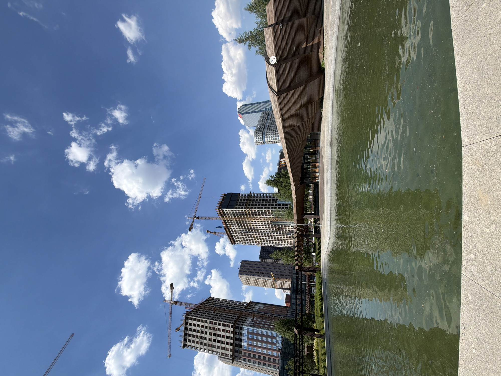 The image depicts a large, outdoor public space with a body of water in the foreground. Behind the water, a unique, wavy wooden structure with a covered seating area is visible. In the background, several tall, modern buildings, some of which are under construction with cranes, dominate the skyline. Green trees and landscaping are present throughout the scene, suggesting a park or urban green space. The sky is blue with scattered white clouds, indicating fair weather. The overall impression is one of urban development and leisure. This scene is located in the city of Moscow, Russia.