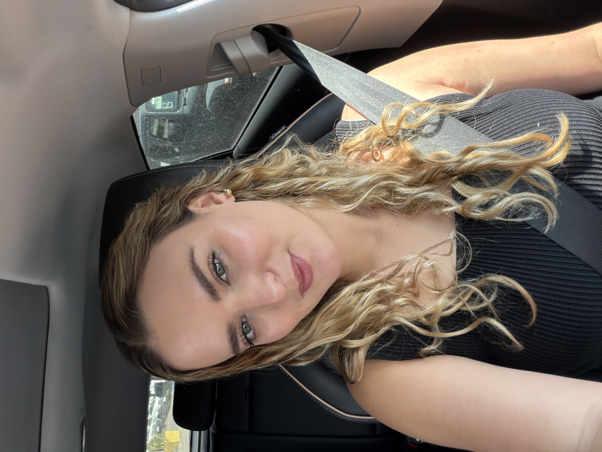 A young woman is taking a selfie while seated in the passenger seat of a car, likely in Puebla, Mexico. She looks directly at the camera with a soft smile, her head tilted to her left. Her long, curly hair, a mix of blonde and light brown with highlights, frames her face and flows over her shoulders. She has striking green eyes and is wearing a black ribbed top and a delicate gold chain necklace. A small gold stud earring is visible on her right ear.
The car's interior is visible, featuring a black leather-like seat with light-colored stitching and a grey ceiling. A black seatbelt crosses her chest and shoulder. Above the car's window, the word "AIRBAG" is clearly visible on the pillar. Bright natural light, suggesting daytime, streams into the car, casting highlights on her hair and skin.
Through the car's window, a blurry outdoor scene is visible, appearing to be a street with reflections and possibly a building or another vehicle in the background. The window itself has some dust or streaks. The overall impression is a casual, relaxed moment captured in transit.