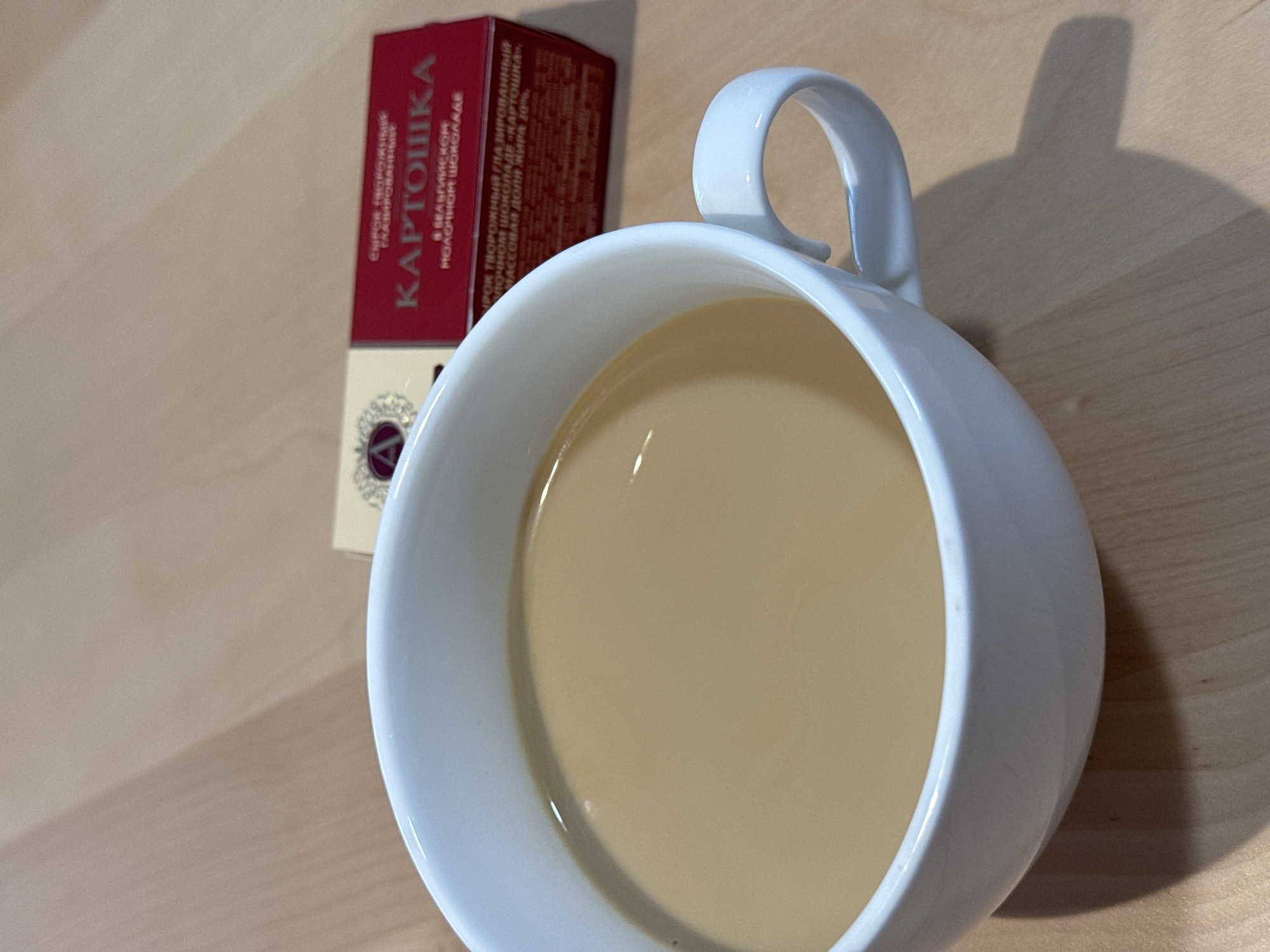 A high-angle shot shows a white ceramic mug filled with a light brown liquid, likely coffee or tea with milk, sitting on a light brown wooden surface. Beside the mug is a small, partially visible red and cream-colored box labeled "KARTOCHKA" in Cyrillic script. The box appears to contain some kind of food item, possibly a confection. A shadow from the mug falls on the wooden surface to the right.