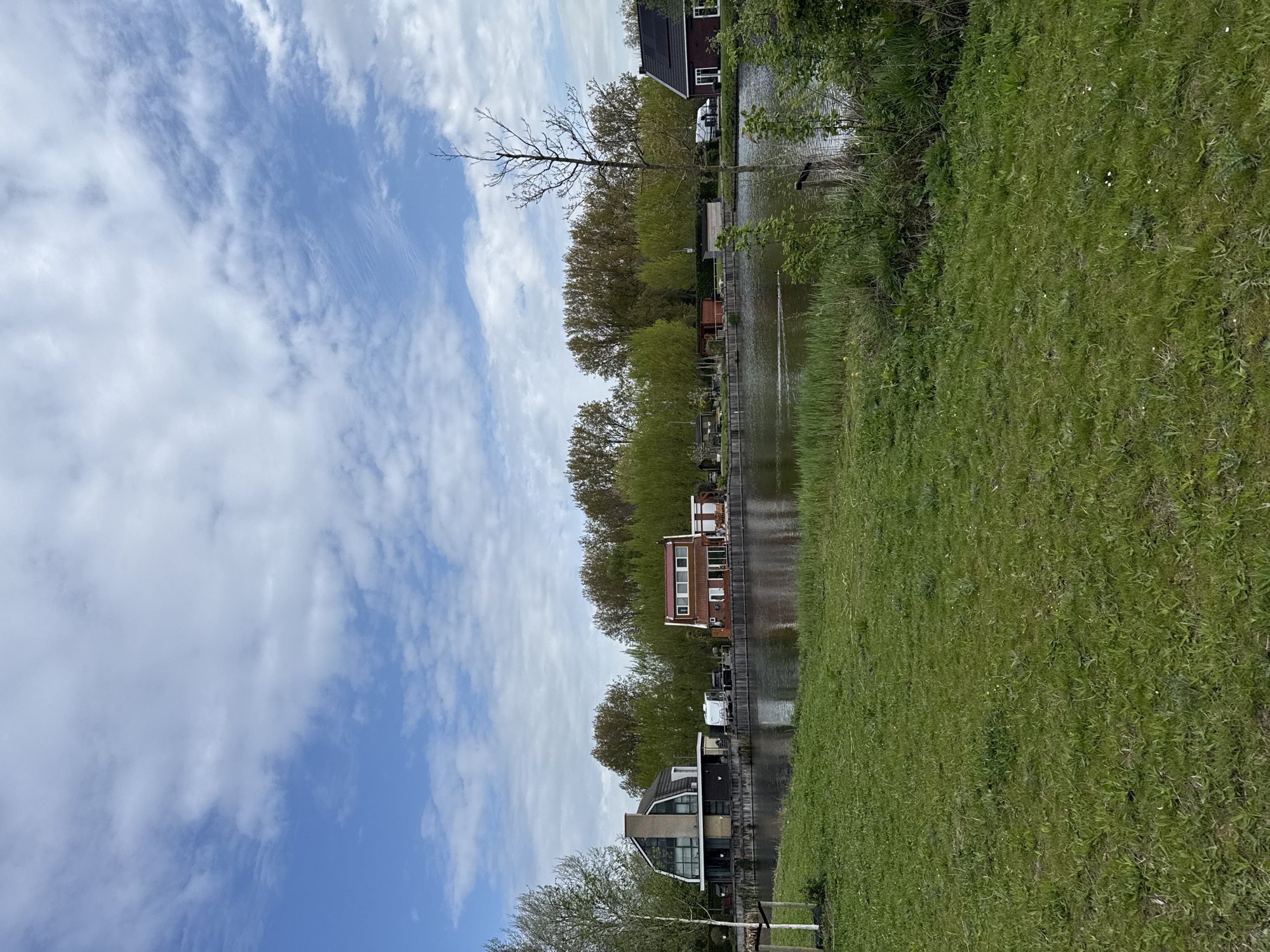 The image displays a serene landscape featuring a waterway, residential buildings, and lush greenery under a partly cloudy sky.
In the foreground, a vibrant expanse of green grass, dotted with small white flowers, occupies the right side of the frame, sloping gently towards a calm body of water. The water, likely a canal or narrow river, runs horizontally across the middle of the scene, reflecting the blue sky and surrounding vegetation.
Along the far bank of the waterway, a line of houses with varied architectural styles is visible. One house features traditional brick construction with a red roof, while another appears more modern with large windows and a dark roof. Some roofs show solar panels. Behind the houses, a dense row of trees, displaying various shades of green foliage from budding to full leaf, creates a natural backdrop. A prominent bare tree with thin branches stands out against the sky near one of the houses. Another smaller, lighter-colored tree is seen in the lower left. Docks or retaining walls line the water's edge in front of the houses, and a white vehicle, possibly a caravan, is parked nearby.
The sky, occupying the upper portion of the image, is a bright blue with scattered, streaky white clouds, indicating fair weather. The scene is illuminated by natural daylight, suggesting it is daytime. There are no people visible, and no specific activities are taking place, contributing to the tranquil and peaceful atmosphere. There is no visible text.