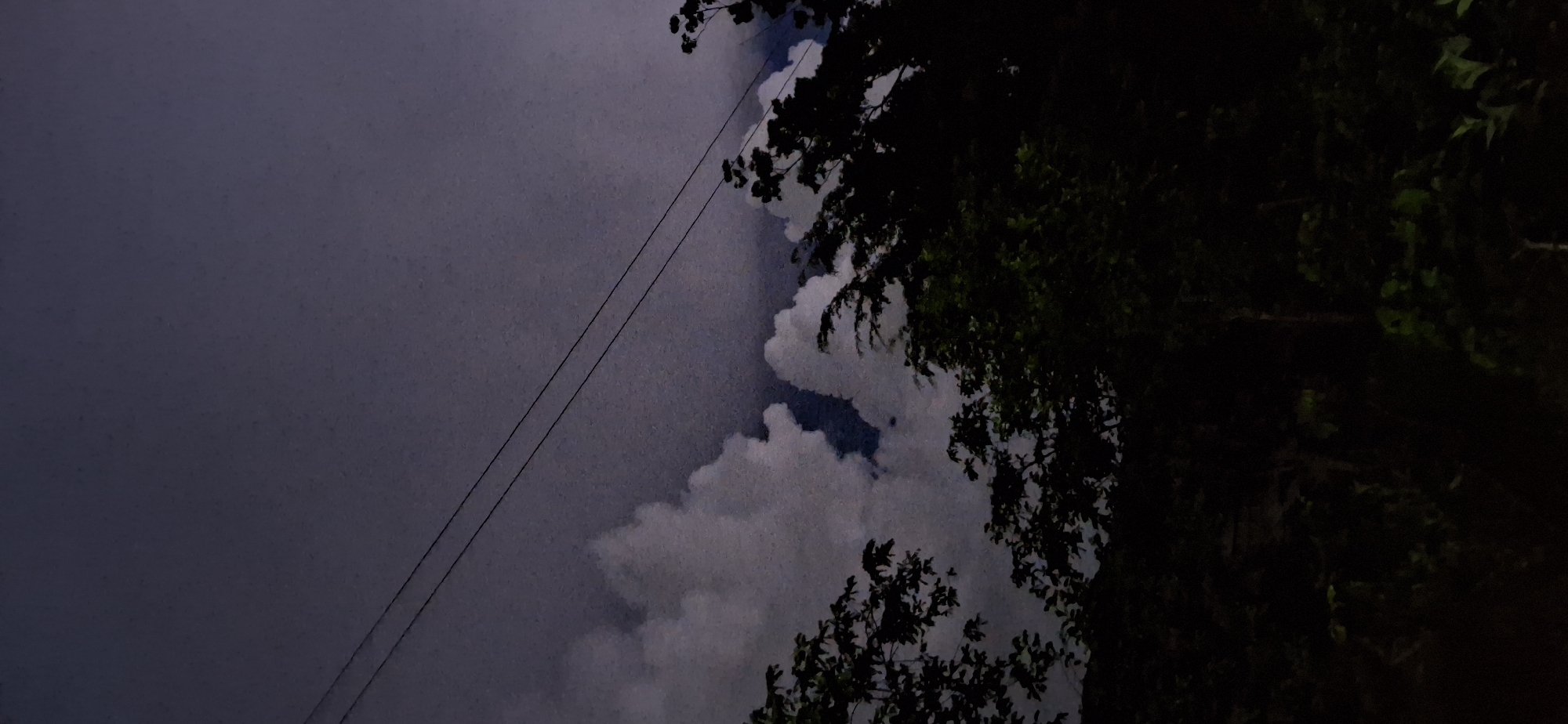 The image depicts a dark, overcast sky over what appears to be a natural landscape, possibly on the outskirts of Vallecito, Venezuela. Two thin power lines are visible, stretching diagonally across the upper portion of the frame. Below the sky, large, white, puffy clouds are interspersed with dark patches, suggesting either breaks in the cloud cover or darker cloud formations. In the foreground, silhouettes of trees and dense foliage are visible, rendered almost black by the low light conditions. The overall impression is one of a twilight or nighttime scene, with limited illumination. There are no people or discernible actions in the image.