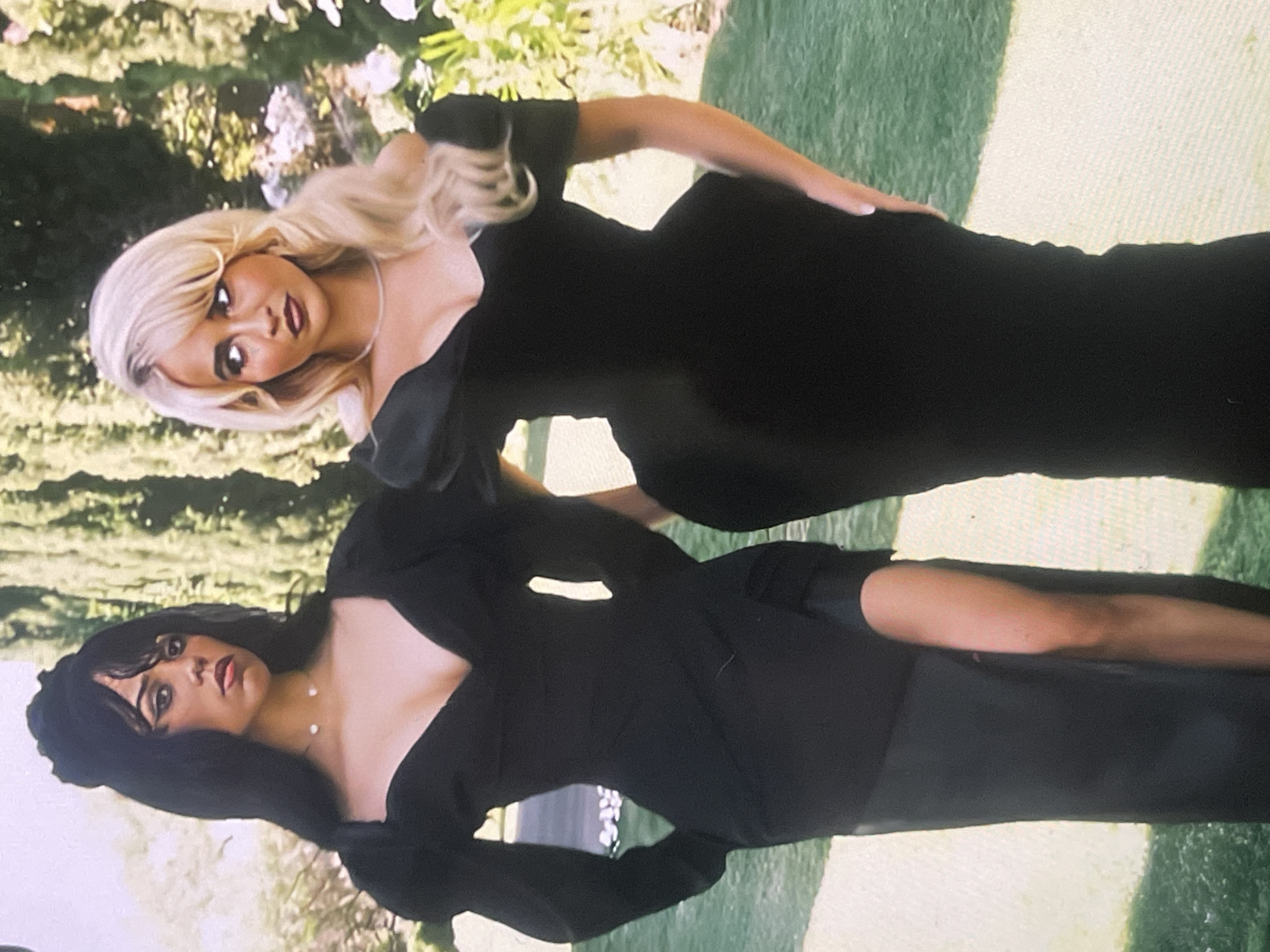 The image displays two women posing outdoors in what appears to be a garden or park setting, possibly in Poltava, Ukraine. The scene is brightly lit by natural daylight, suggesting a clear day.
On the left side of the frame, a woman with long, dark hair styled with bangs looks directly at the camera with a focused, serious expression. She is wearing an elegant black dress featuring a deep V-neckline and distinctive puffy sleeves. A delicate necklace with two small white pearls is visible around her neck. Her makeup includes dark eyeliner and a rich, deep red lipstick.
To her right stands another woman, characterized by voluminous, wavy blonde hair. She also looks directly at the camera, maintaining a serious or sultry gaze. She is dressed in a sleek, full-length black gown with short sleeves. A thin, light-colored necklace adorns her neck. Her right hand rests gracefully on her hip.
Both women are wearing formal black attire, suggesting they are attending or posing for a special event or photoshoot. The background is softly blurred, showcasing vibrant green grass, interspersed with various trees and leafy foliage, and hints of a lighter-colored pathway or ground. There is no visible text in the media file.