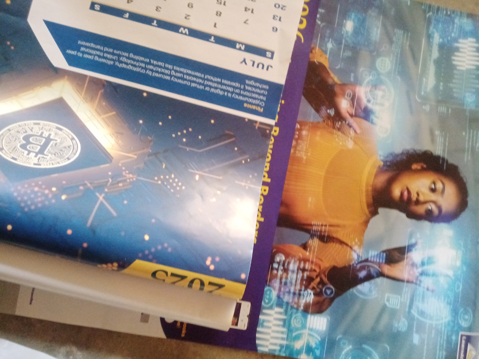 The image shows a close-up, slightly angled view of a poster or calendar. The poster features a woman in a yellow shirt interacting with a holographic interface that displays various data and icons. To the left, a portion of what appears to be a Bitcoin symbol is visible, superimposed on a blue background with scattered lights, resembling a city skyline. At the bottom left, the year "2023" is clearly visible in large yellow font. Text on the poster includes the word "Finance" and the month "JULY". The image does not contain any identifiable landmarks or context that would suggest a specific location in Jalingo, Nigeria. The scene depicts a technological and financial theme.