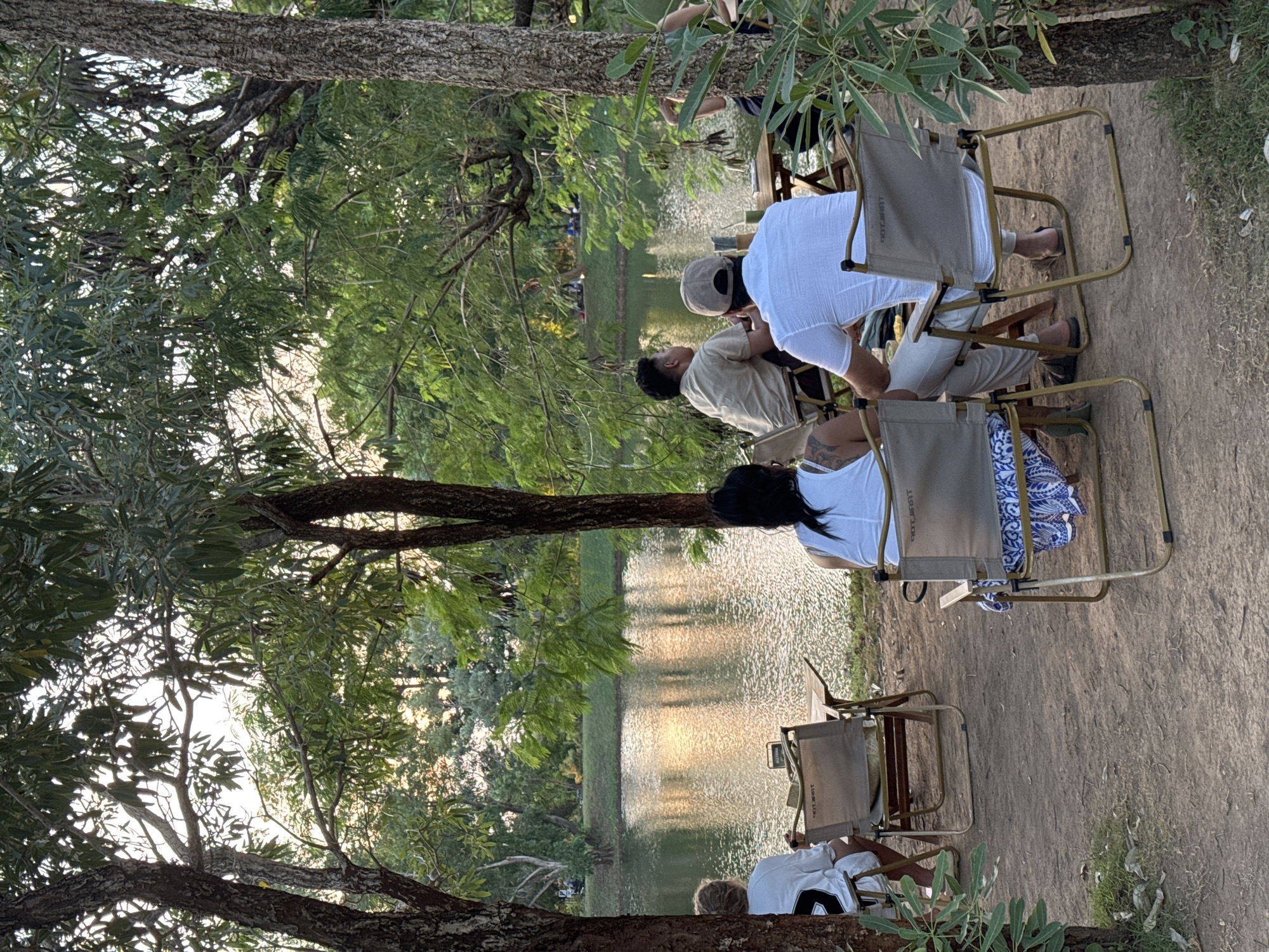 The image depicts a group of people relaxing on folding chairs by a body of water, likely a lake or pond, in an outdoor setting. The location is suspected to be in Pecatu, Indonesia. Several individuals are seated, facing the water. The area is shaded by trees, which frame the view and reflect in the water. There's a natural ground surface visible where the chairs are placed. People are seated in chairs. One is wearing a white t-shirt, another a brown shirt.
