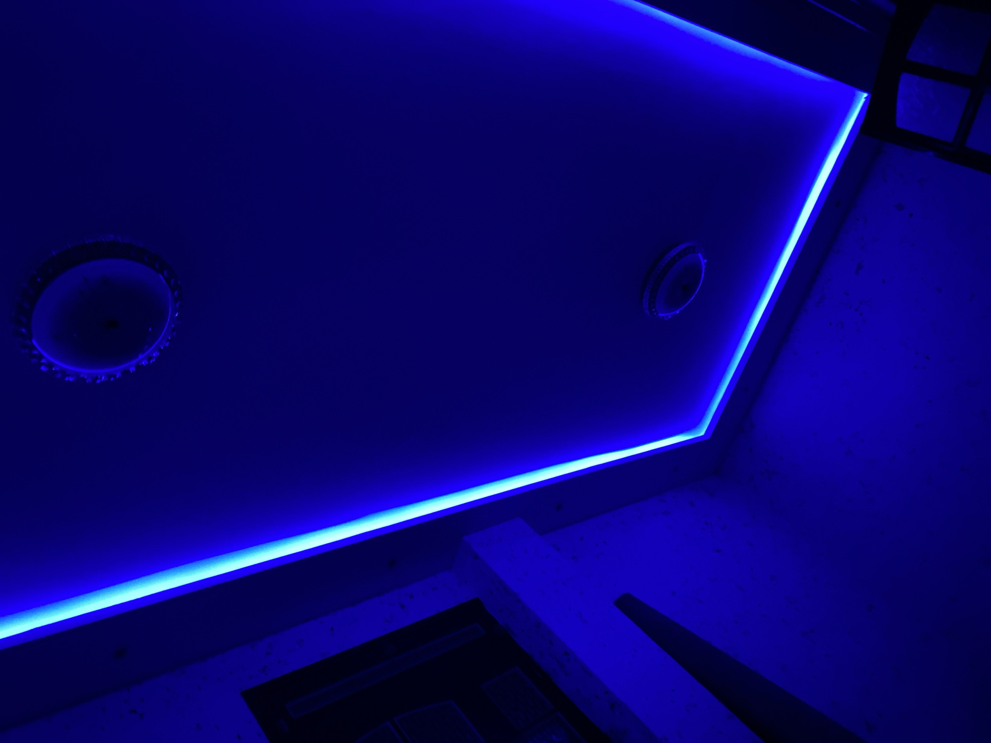 The image depicts a room interior illuminated by blue LED strip lighting installed around the ceiling perimeter, creating a strong blue cast. Two circular recessed lights are visible on the ceiling. The walls appear to be light-colored with a speckled texture. A partial view of some dark, angular furniture or architectural elements is visible in the lower portion of the image. The overall impression is a modern, stylized space with a cool, artificial lighting scheme. There is no indication in the image that can suggest the location in the city of Baraki, Algeria.