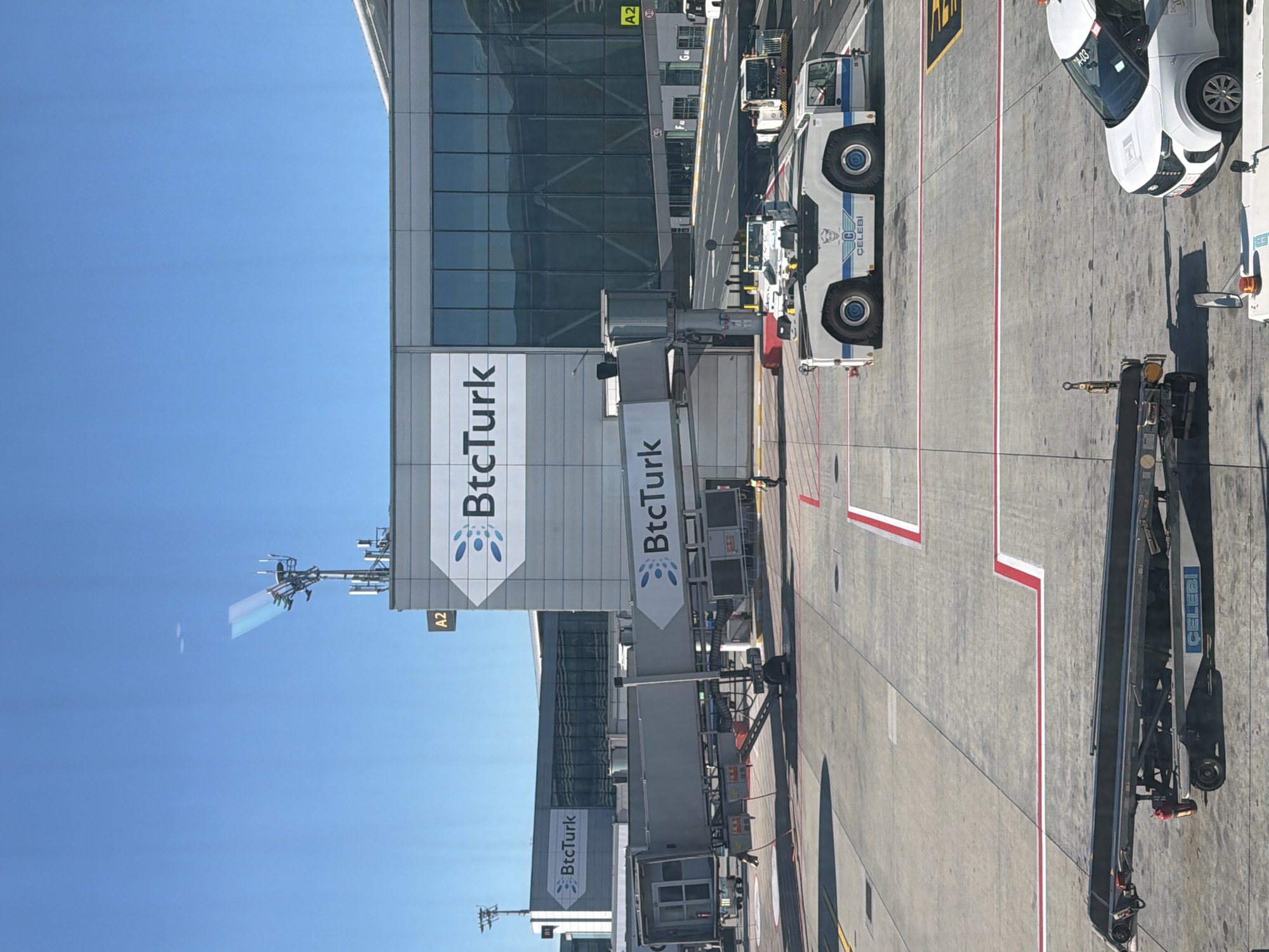 The image shows an airport tarmac in Arnavutköy, Turkey, on a clear, sunny day. A jet bridge, labeled with "BtcTurk," connects to a terminal building also bearing the "BtcTurk" logo. Two airport tow tractors are visible on the tarmac. One is a white tractor with blue accents and the "Çelebi" logo, positioned in the foreground. Another similar tractor is further back. A baggage conveyor belt is seen on the left side of the frame, with a fire extinguisher attached. A white passenger vehicle with "A-03" markings is parked on the right. The tarmac is marked with red and white lines. In the background, the modern terminal building features large glass windows, and a communication tower with antennas is present. A single individual wearing an orange vest is walking on the tarmac near the jet bridge.