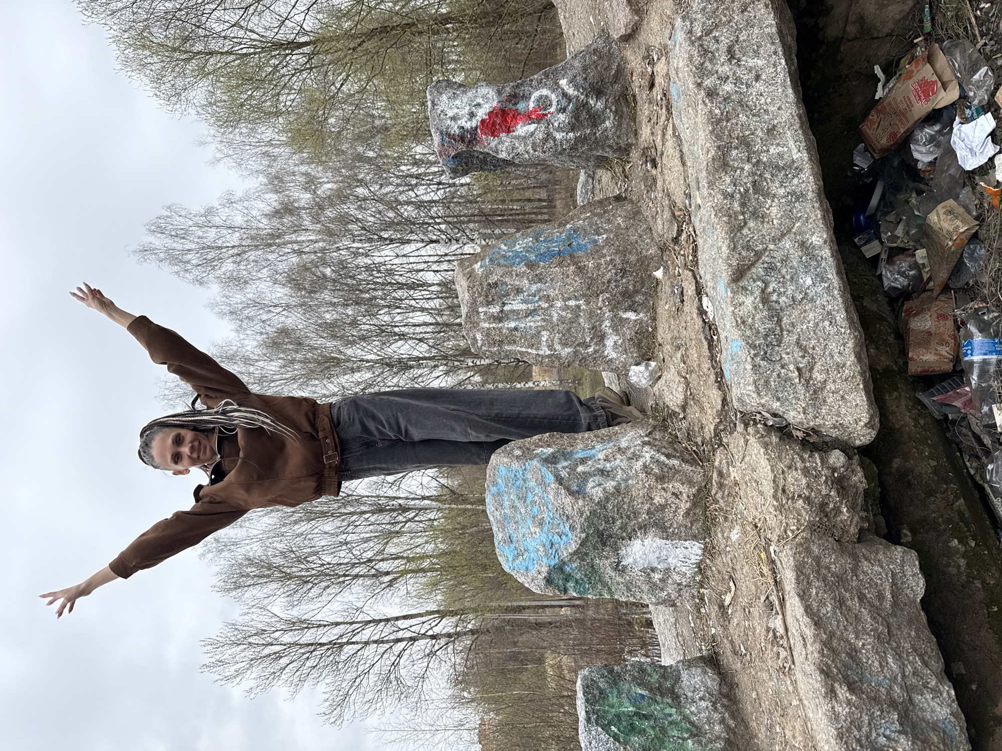 A young woman with long, light-colored braided hair is standing on large, gray rocks in Rusanovo, Russia. She is wearing a brown jacket and dark gray pants, and she is smiling with her arms outstretched. The rocks on which she is standing are painted with graffiti in red, blue, and green. Behind the rocks is a stand of trees with bare branches. To the right of the rocks is a pile of garbage consisting of food containers, plastic bottles, and other debris. The sky is overcast.