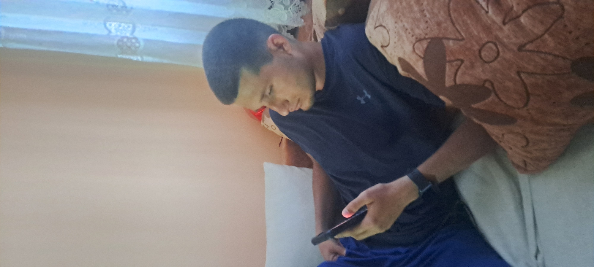 A young man is lying on a bed, looking at a phone. He is wearing a dark blue t-shirt and blue athletic shorts, and a smart watch is visible on his left wrist. He is resting against a patterned brown pillow and a white pillow. A curtain is visible behind him, and a beige wall is to his left. There is no indication of Burgas, Bulgaria in the image.