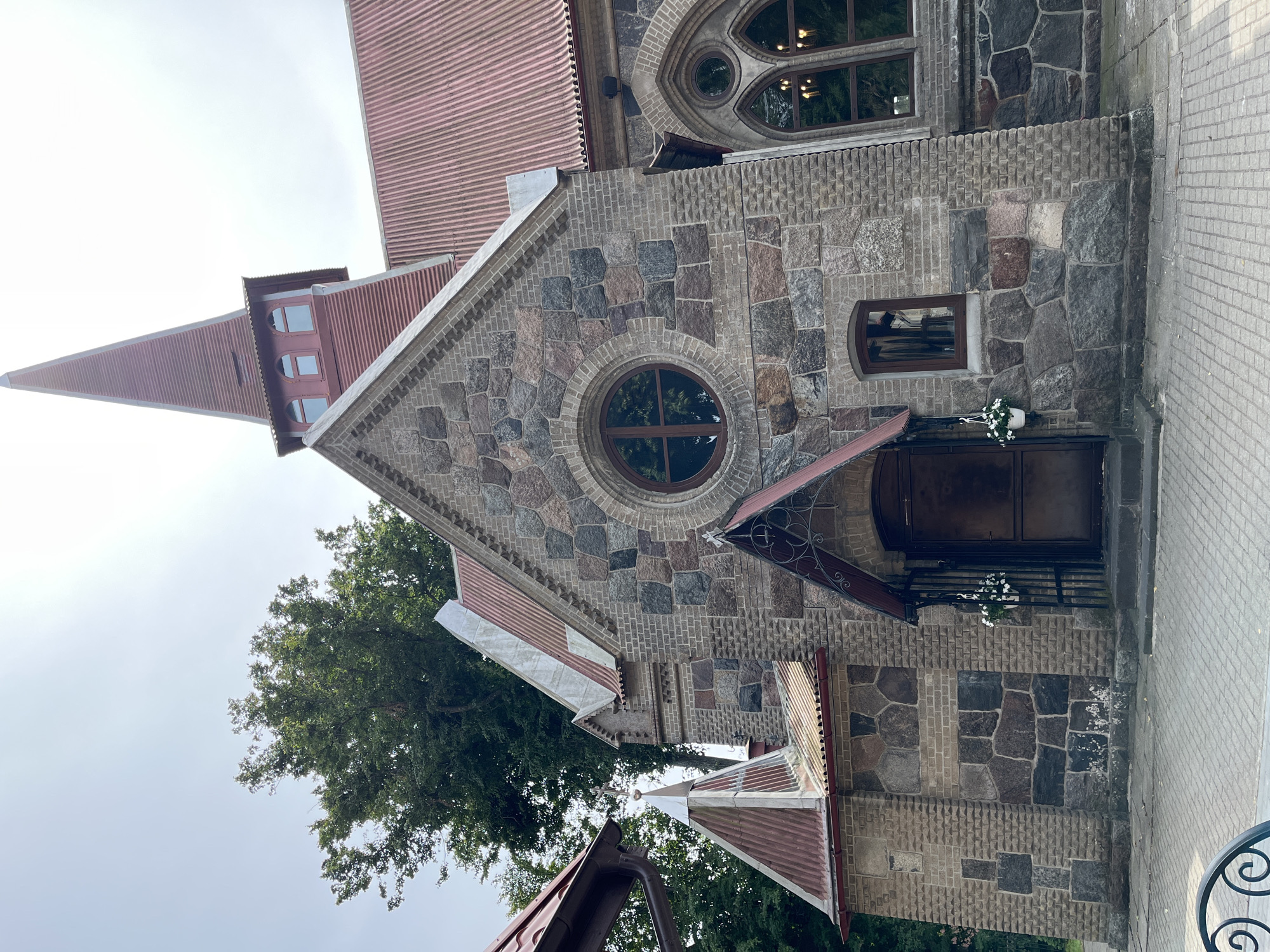 The image shows the exterior of a stone and brick church in Yantarny, Russia. The church has a prominent red, corrugated metal roof and a tall, pointed steeple. The main facade is constructed of large, irregularly shaped stones in various shades of grey, brown, and red, interspersed with sections of lighter brickwork. A circular window with wooden framing is centrally located on the upper part of the facade. Below this, an arched doorway is covered by a dark metal gate with decorative scrollwork. A small window with a dark wooden frame is situated to the right of the doorway. To the right of the main facade, a section of the church features Gothic-style arched windows, one of which is visible and shows reflections of the surrounding trees and interior lights. White potted flowers hang on either side of the entrance gate. The church is surrounded by trees and is situated on a paved courtyard. The sky is overcast.