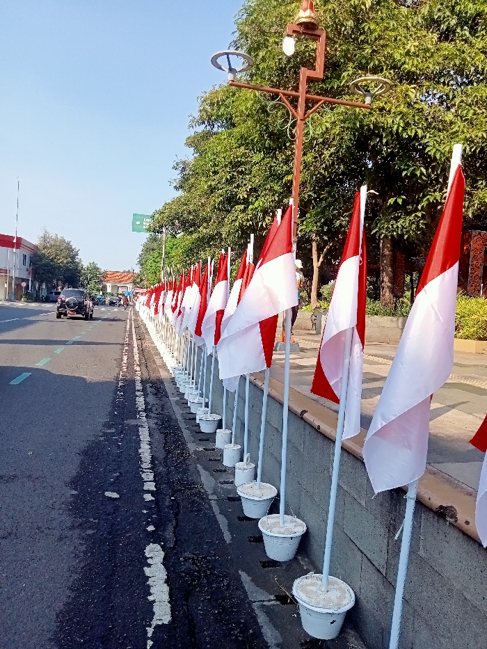 The image depicts a roadside scene in Cikarang, Indonesia, under a clear blue sky. A long row of approximately 30-40 Indonesian national flags (red over white horizontal bicolour) is prominently displayed on white poles along the right side of an asphalt road. Each flag pole is anchored in a white bucket filled with a solid substance, placed on a raised concrete curb or median.
The road, visible on the left, features a dashed white line and a faint dashed green line. A dark-colored SUV (appearing to be a Jeep Wrangler) travels away from the camera in the left lane. Other vehicles are faintly visible in the far distance on the road.
On the right side, beyond the flag display, a landscaped area with dense green trees and bushes is present. A decorative, dark brown streetlamp with two circular light fixtures and ornate metalwork stands upright in this area. Further down the row of flags, another similar streetlamp is partially visible. A green rectangular street sign with white text is discernible in the far distance on the left side of the road, reading "Jawa Barat" (West Java).
Two individuals are observable in the distance on the right, walking alongside the flag display. One figure, closer to the camera, appears to be wearing a light-colored top and dark trousers, with a light object (possibly a helmet or hat) on their head. The scene is illuminated by direct sunlight, casting minimal shadows.
