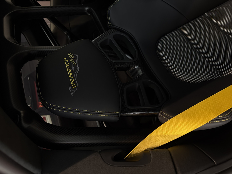 The image presents a detailed view of the interior of a high-performance vehicle, specifically focusing on the passenger seat area. The seat is a black racing-style bucket seat, constructed with black leather and yellow contrast stitching along its seams. The headrest features an embroidered yellow "WEISSACH RS" logo, depicting a stylized racetrack outline. Below the headrest, the seatback incorporates multiple openings and is accented with carbon fiber elements. The seat's base includes a textured, possibly mesh, black fabric insert. A prominent bright yellow seatbelt is visible, extending diagonally from a retention point on the left side of the image towards the lower right. Behind the seat, a black carbon fiber roll cage structure is visible, forming a protective frame within the vehicle's cabin. The immediate background outside the car window is dark and out of focus, offering no discernible details of an exterior environment. No individuals are present in the scene. Based solely on the visual information provided, a specific location within the city of Regensburg, Germany, cannot be determined.