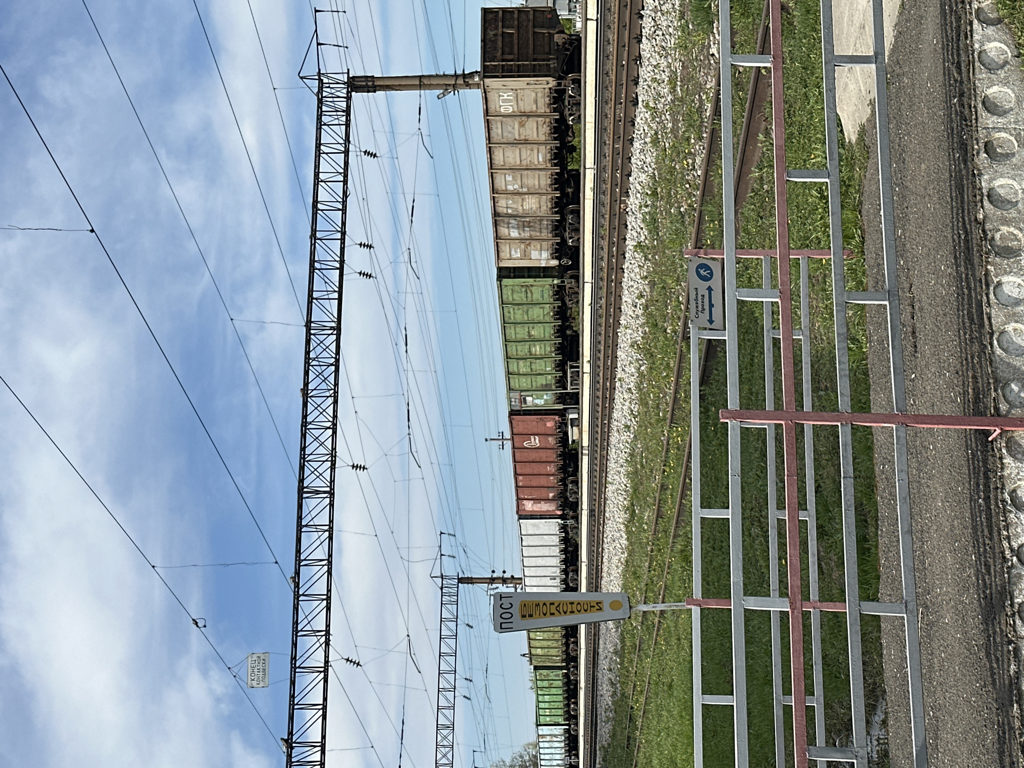 The image shows a railway scene in Кечёво, Russia. A train consisting of multiple freight cars of varying colors (brown, green, and white) is on the tracks. Overhead, there are power lines supported by a metal structure. A pedestrian crossing with a metal barrier and signage is in the foreground. One sign reads "POST" vertically. The other is a directional sign for crossing. The sky is visible in the background with scattered clouds.