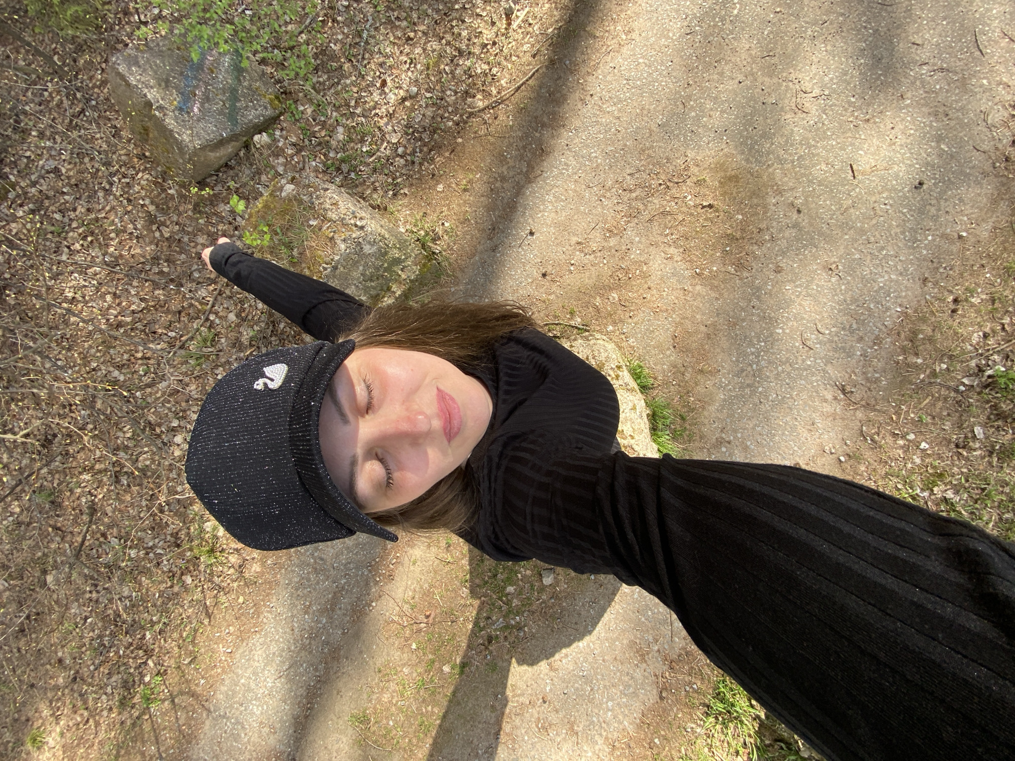 A woman with fair skin is wearing a black, long-sleeved, ribbed dress and a black cap with a silver swan emblem. She is lying sideways on the ground, possibly on a grassy area adjacent to a concrete path. One arm is extended towards a large concrete block partially covered in moss, which is adjacent to foliage and twigs. The ground consists of a concrete path and areas of dirt and leaf litter. The photo appears to be taken outdoors in daylight with shadows present. The location is Noviy, Russia.
