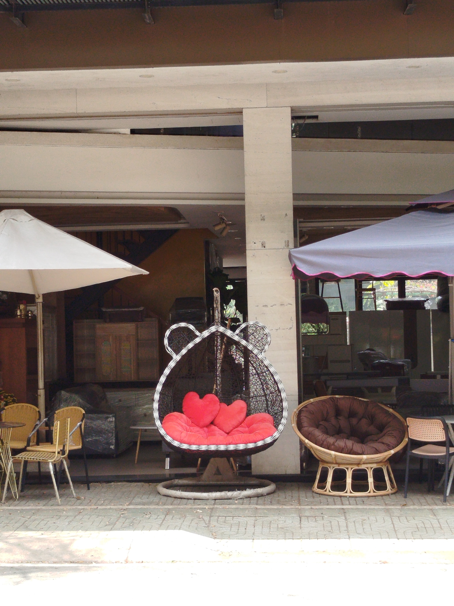 The image shows a storefront in Liên Nghĩa, Vietnam. Various pieces of outdoor furniture are displayed on the sidewalk. These include a hanging wicker chair with red heart-shaped cushions, a brown wicker papasan chair with a round cushion, and several yellow wicker chairs with a small table. Two umbrellas provide shade: a white one on the left and a grey one on the right. Through the storefront windows, the interior of the business is visible, showing stacked furniture and interior design elements, including a staircase. The storefront is supported by a large white pillar. The sidewalk is tiled and slightly dirty.