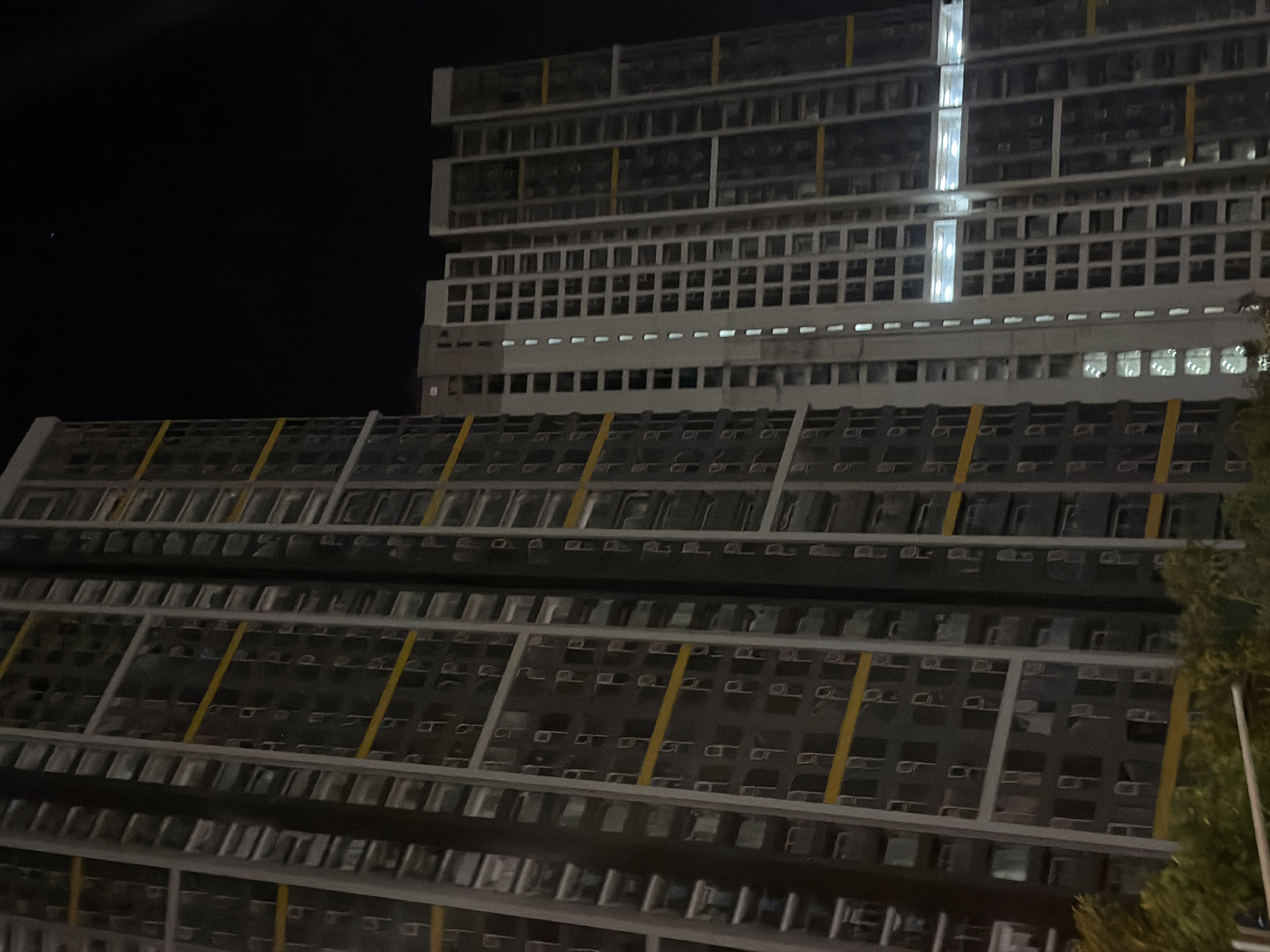 The image captures a nighttime view of a large, modern building in Đà Nẵng, Vietnam. The building is multi-story, with a distinctive grid-like facade. Rows upon rows of modular units or windows are visible, many appearing dark, suggesting an unoccupied or nighttime state. Some sections of the building have illuminated windows, casting a soft glow.
A prominent feature is the staggered design of the building, with upper sections projecting out over lower ones. Yellow diagonal lines are subtly integrated into the facade of the lower sections, adding a touch of visual interest. The upper part of the building is taller and more rectilinear, featuring a prominent vertical strip of bright, cool-toned illumination.
The scene is set against a dark, clear night sky. To the right of the building, the dark foliage of a tree is visible, providing a contrast in texture and form to the sharp lines of the architecture. The overall impression is one of urban architecture at night, with a focus on the geometric patterns and the interplay of light and shadow. There is no visible text in the image.