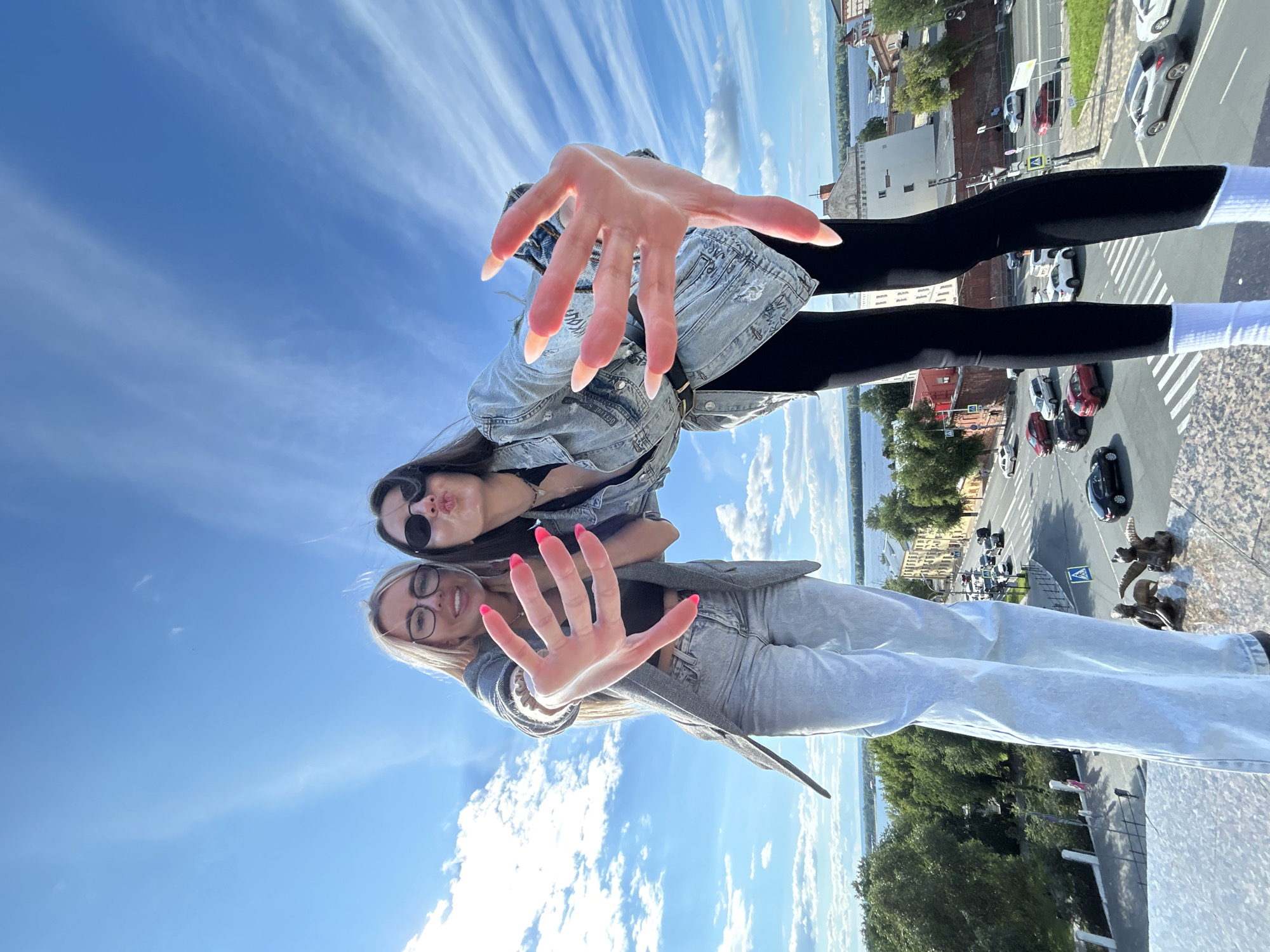 This is a low-angle, wide-shot photograph taken outdoors on a clear, sunny day. The sky is a vibrant blue with wispy white clouds. The image is oriented such that the ground appears above and the sky below.
In the foreground, two young women are captured in a selfie. The woman in the upper part of the frame is leaning forward, her left hand with long, light-colored nails extended towards the camera, appearing very large due to the perspective. She wears a denim jacket over a dark top and black leggings with white socks. She is wearing sunglasses and has her lips pursed.
The second woman is positioned below the first, her face and upper body visible. She wears glasses and a denim jacket over a dark top and light-colored jeans. Her right hand is also extended towards the camera, with bright pink nails. Both women appear cheerful and playful.
The background reveals a cityscape with buildings, trees, and a road with cars. The perspective suggests they are standing on a high vantage point, possibly a rooftop or balcony. The presence of buildings and a body of water in the distance indicates an urban or semi-urban setting. The time of day is clearly daytime, evidenced by the bright sunlight and clear sky. There is no visible text in the image.