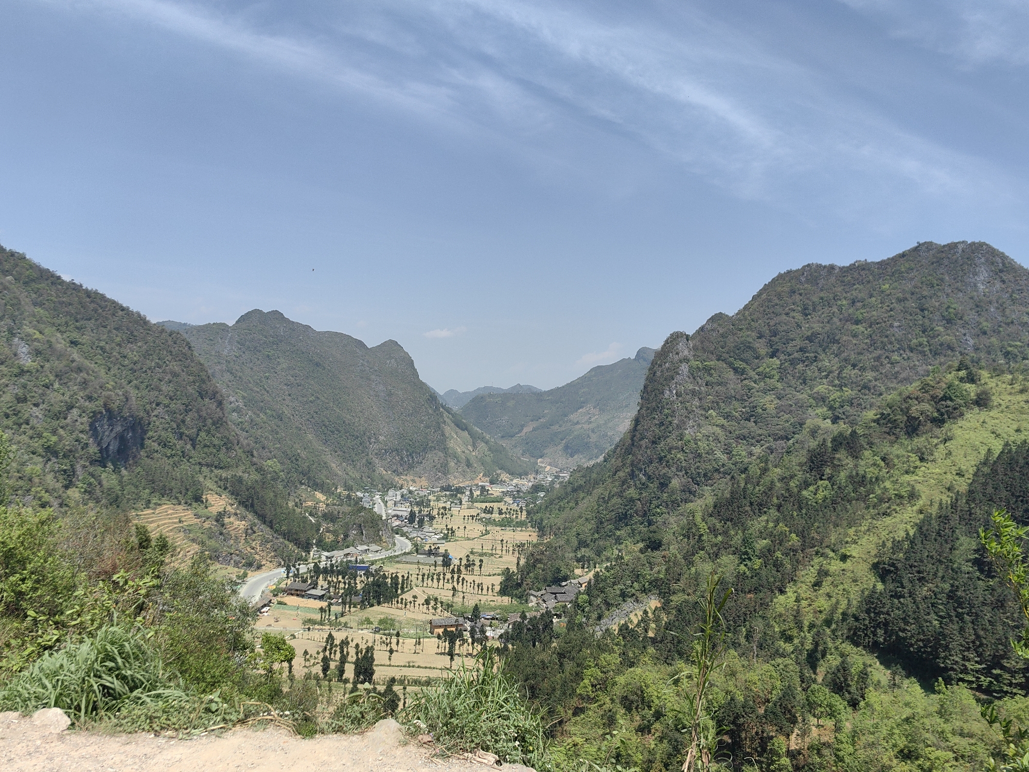 The image is a wide, daytime shot of a mountainous valley, likely in the Sà Phìn Commune, Vietnam. Two prominent, forest-covered mountain ranges frame a valley containing a town or village with houses and open fields. The sky is pale blue with faint, wispy clouds. The fields appear to be cultivated, with rows of trees punctuating the landscape. A road winds its way through the lower valley and into the town. The foreground shows green vegetation and a dirt path or hillside. The overall scene depicts a rural setting in a mountainous region.