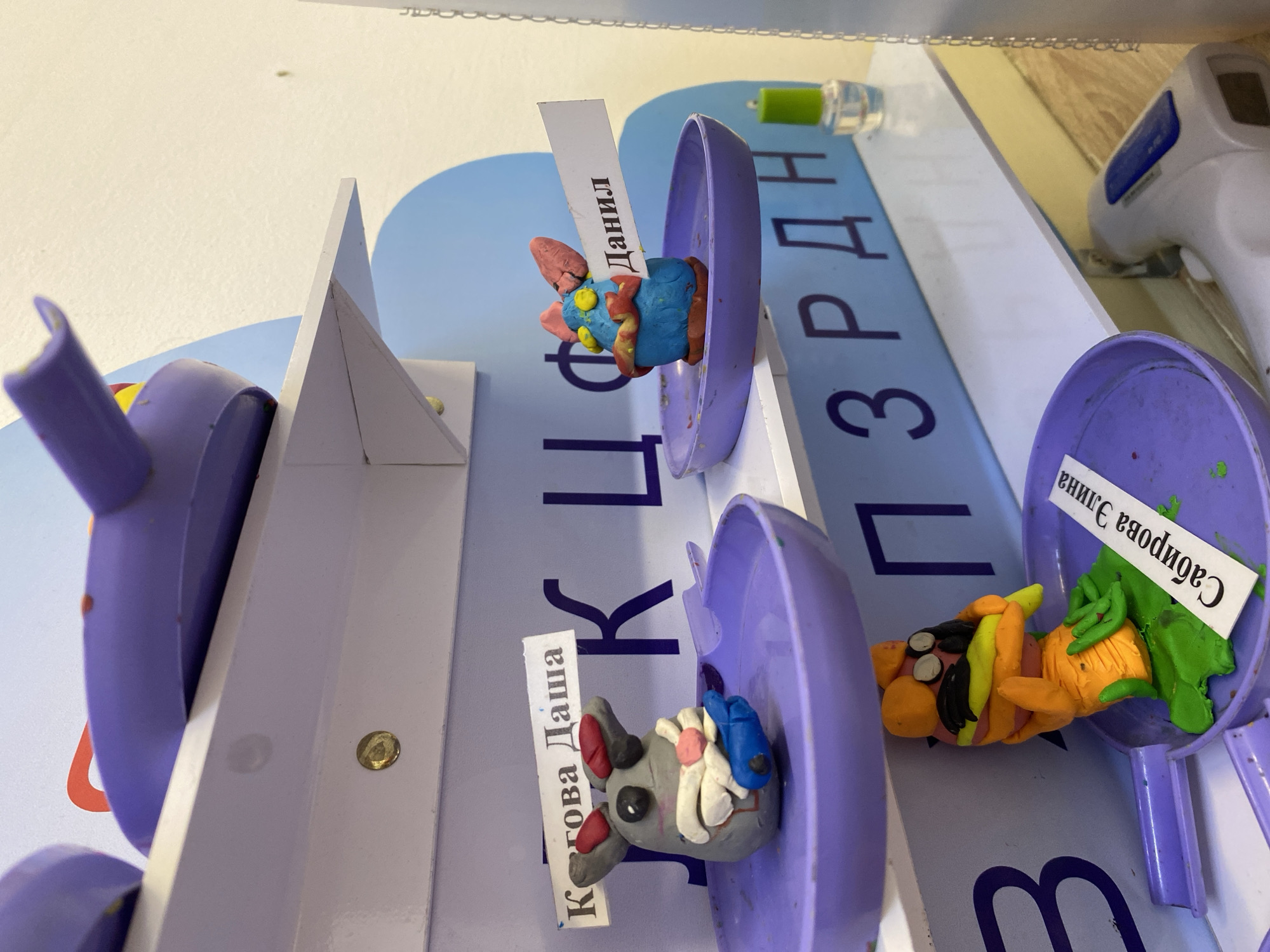 The image presents a close-up shot of a child's artwork display, likely within a school or daycare setting. Three plasticine clay figures are positioned on a stepped shelf. Each figure is housed within a repurposed plastic food container, painted purple. Next to each figure is a small paper label with Russian writing, presumably identifying the name of the child who created the artwork.
The figure on the left is predominantly grey, featuring some pink and white details. The central figure is mainly blue and red. The figure on the right is a combination of orange, yellow, green, and black. The backdrop appears to be a board with printed Russian text in large purple letters. A non-contact thermometer and a vial of liquid are visible in the background.
There is no contextual information to confirm the location to be in Noviy, Russia.