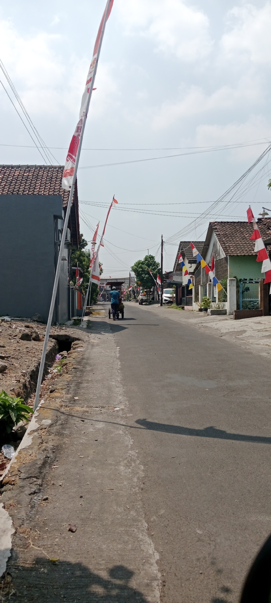 The image depicts a street scene in Maguwoharjo, Indonesia, on a clear, sunny day. The street is paved and appears to be a residential area, with houses lining both sides. Red and white Indonesian flags are prominently displayed along the street, suggesting a national celebration or event.
On the left side of the street, a dark gray house with a tiled roof is visible. A drainage ditch runs along the edge of the sidewalk in front of this house, containing some debris. A tall, thin white pole with a flag attached stands near the foreground.
On the right side of the street, several houses with tiled roofs are visible. These houses have varying colors, including light blue and green. More Indonesian flags are mounted on poles in front of these residences.
In the middle of the street, a person wearing a blue shirt is pushing a two-wheeled cart. The cart appears to be carrying some items. The person is moving away from the camera, down the street. Further down the street, two cars are parked or moving, one white and one darker colored. Overhead power lines crisscross the sky. The overall impression is a quiet, ordinary day in a residential neighborhood adorned for a patriotic occasion.