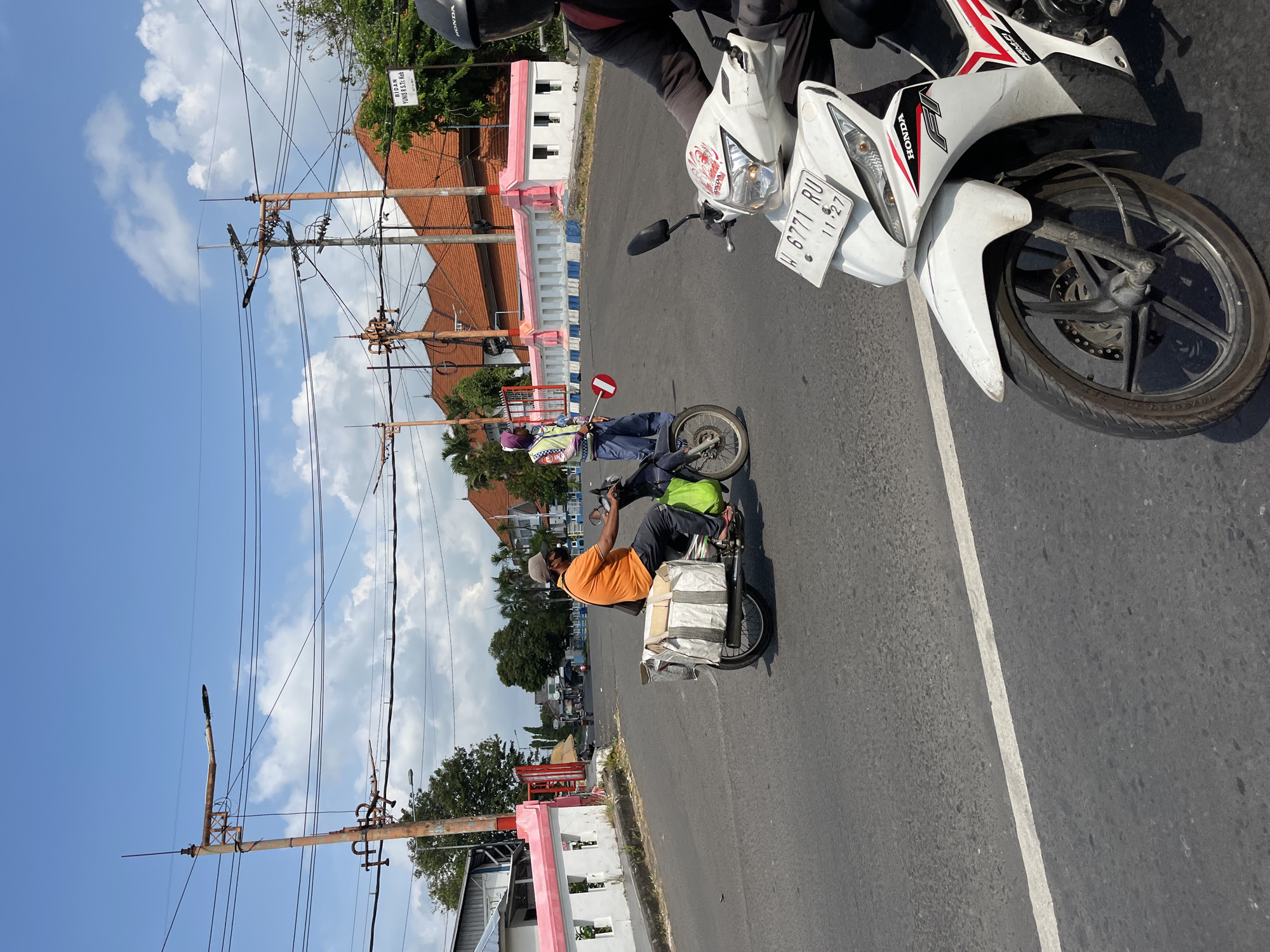 In Kludan, Indonesia, on a sunny day with a clear blue sky and scattered clouds, a street scene unfolds. The road, marked with a white line, features several motorcycles. In the foreground, a white Honda motorcycle, with license plate W 6771 RU, is prominently displayed, its front wheel and part of the body visible. To the left, another motorcycle is seen carrying a rider in an orange shirt, dark pants, and a cap, with a large bag strapped to the back. Further down the road, a traffic officer in a vest and helmet is directing traffic with a stop sign. Behind the officer, a building with a pink and white facade and terracotta tiled roofs stands. Overhead, electrical poles and wires crisscross the sky, common infrastructure in urban areas. A sign that reads "BIDAN YUNIS R. S. Tg. Keb" is visible on a pole. The overall environment suggests a typical day in a bustling Indonesian town.