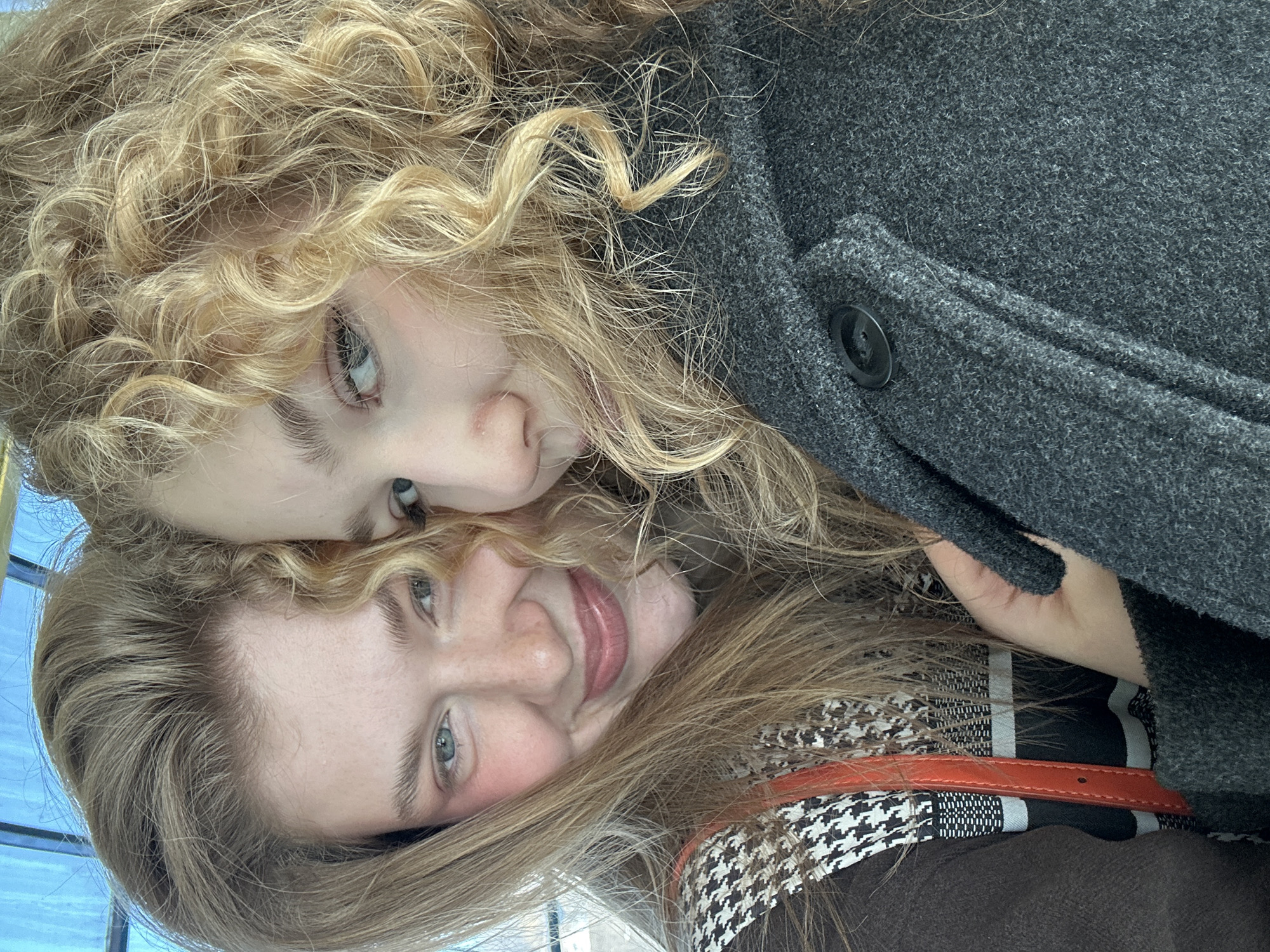 A close-up selfie-style image displays two young adult females in the city of Vorob'evka, Russia. The subject on the left exhibits straight light brown hair, light-colored eyes, and blush on her cheeks, with a slight smile. She wears a dark brown garment, a black and white houndstooth pattern layer, and an orange strap. The subject on the right possesses voluminous blonde curly hair and light-colored eyes, with her gaze directed to the viewer's right. She is dressed in a dark grey textured woolen coat, which includes a visible dark button and a lapel. A hand is partially visible, likely belonging to the right subject, positioned centrally. The background is blurred, showing soft blue and white hues, consistent with an outdoor or brightly lit indoor environment.