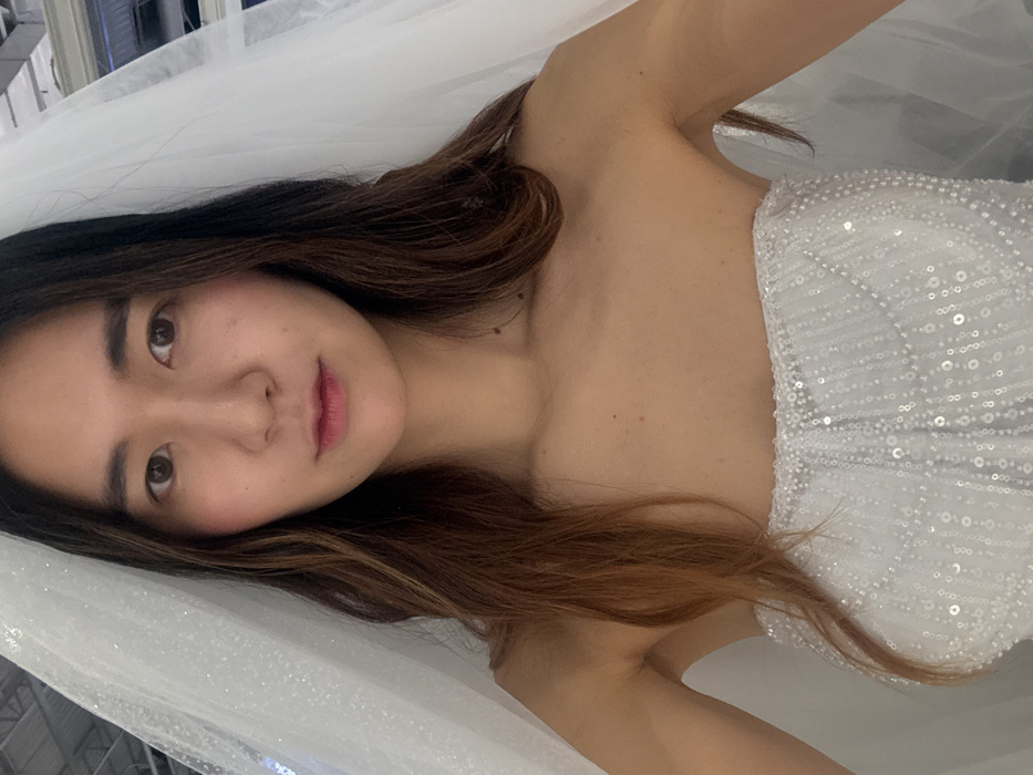 A woman poses in a strapless white dress adorned with reflective sequins. Her dark brown hair, lightened with auburn highlights, cascades over her shoulder. She looks directly at the camera, her expression neutral. A sheer, sparkling white fabric drapes behind her, suggestive of a veil or gown overlay. The setting appears to be indoors, possibly a bridal boutique.