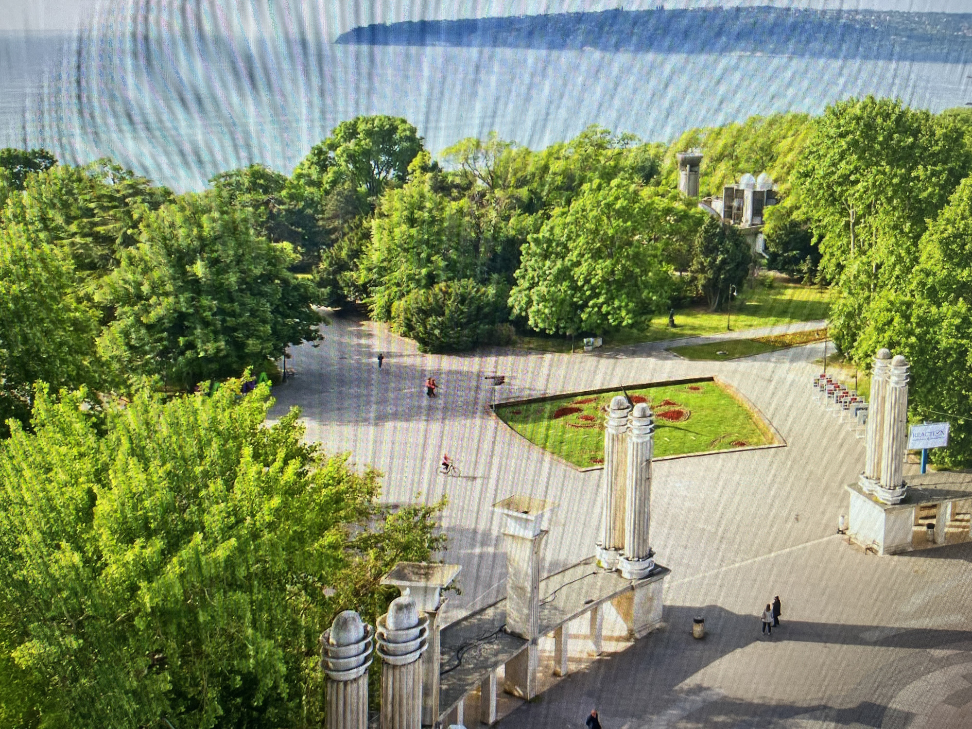 This is an aerial, eye-level view of a park and waterfront area in Varna, Bulgaria. The background features a wide body of water, likely the Black Sea, with a distant, dark-colored landmass. In the foreground, lush green trees dominate the landscape, with a paved pathway winding through the park. Several people are visible: one person is riding a bicycle on the pathway, and a couple walks together. Architectural elements, including ornate columns and a building with multiple domed structures, are scattered throughout the park. A red and white sign with the word "REACTION" is mounted on a post. The scene appears to be a public park, possibly a recreational area by the sea.