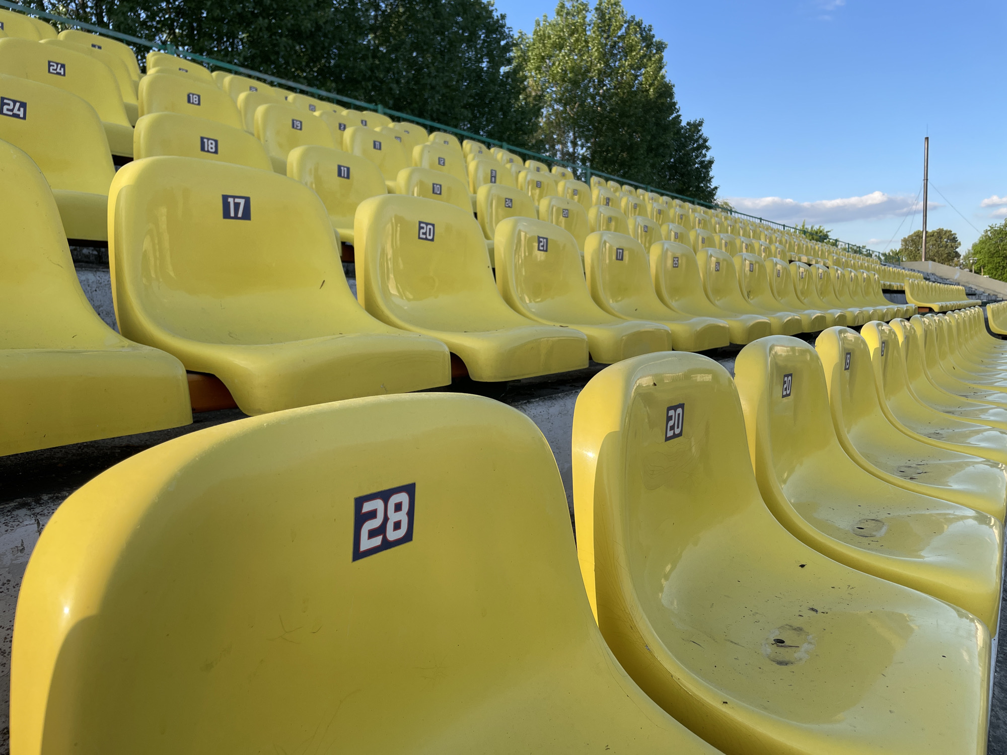 The image shows rows of yellow stadium seats, likely outdoors, with a numbered labeling system, with numbers such as 28, 20, 17, 18, and 24. The seats are curved and appear to be made of plastic. The rows ascend upwards towards the background, where trees and a blue sky are visible. The scene is well-lit, suggesting it is daytime. It is likely a section of a stadium in Brest, Belarus.