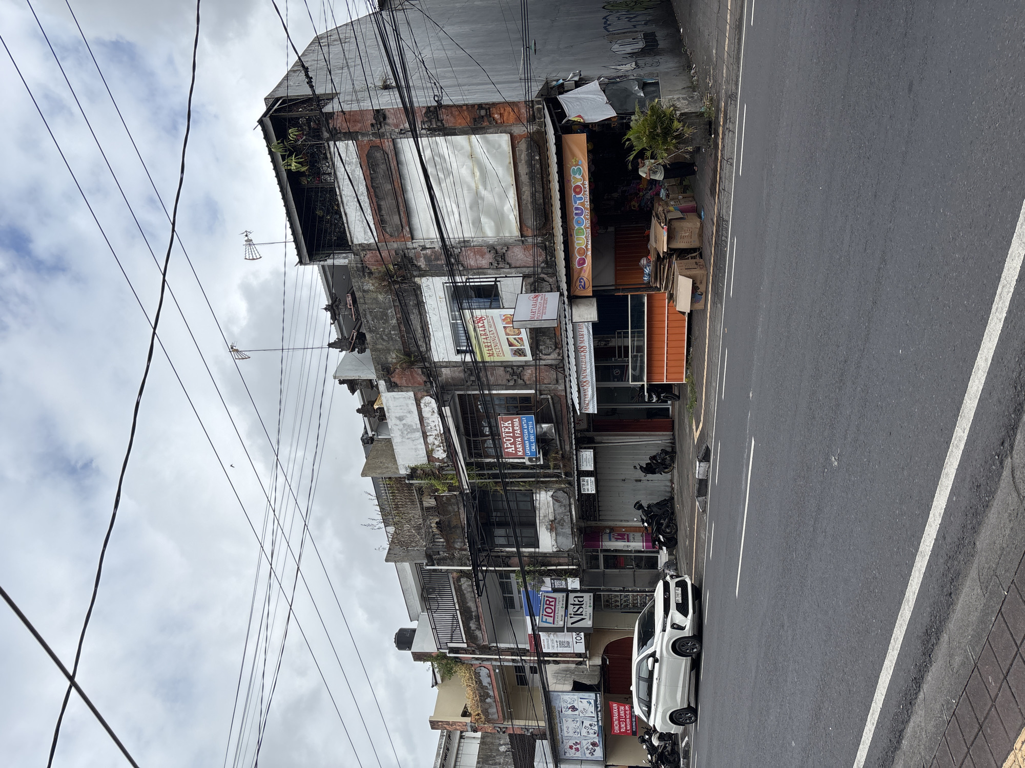 The image depicts a street scene in Denpasar, Indonesia, characterized by a row of buildings, a paved road, and a cloudy sky. The buildings are multi-story structures, some appearing aged with peeling paint and exposed brickwork, while others seem to be more modern. Various businesses are identifiable, including a pharmacy ("APOTEK KARYA FARMA"), a toy store ("DOUBDOU TOYS"), and what appears to be a food establishment ("MARTABAK88 PECENONGEN PREMIUM"). A white car is parked on the left side of the road, and several motorcycles are lined up in front of the shops. A person is visible near the toy store, engaged in an unspecified activity. Electrical wires are prominently strung across the upper portion of the image, and some antennas are visible on the rooftops. The environment suggests a bustling urban area with a mix of commercial activity and residential spaces.