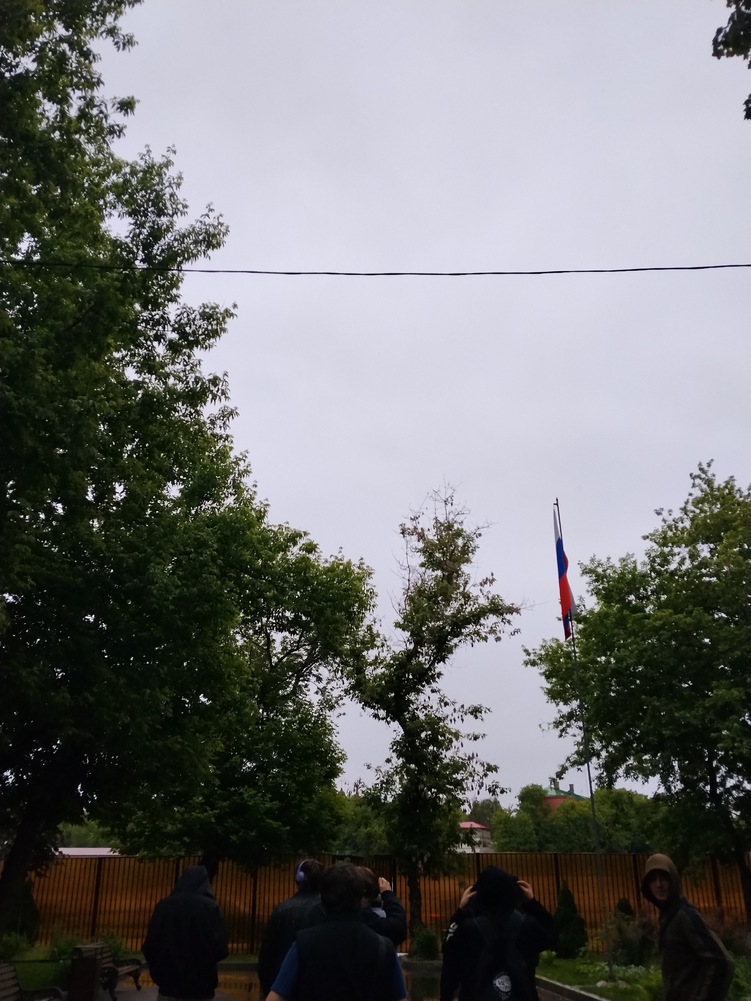 The image shows a group of people standing near a wooden fence, likely on a sidewalk or park path, with trees lining the area. In the background, a Russian flag is visible flying from a pole, positioned between two trees. The sky is overcast. The scene is set in a public space, possibly a park or street with a view of a government building or embassy. Based on the presence of the Russian flag and the architectural styles possibly seen behind it, this image may be located in Moscow, Russia.
