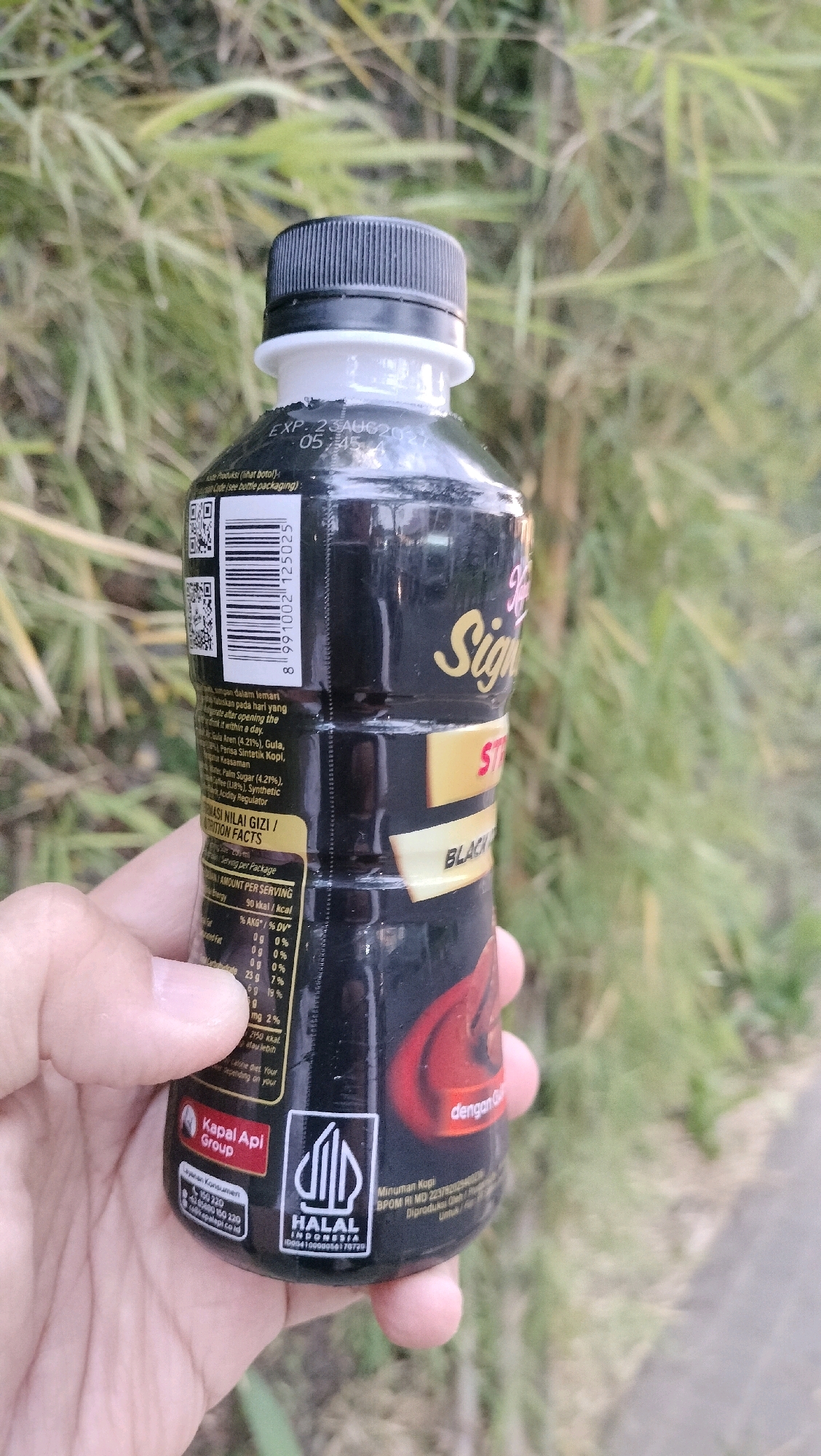 A hand holds a black plastic bottle of "Signature Black Coffee" in an outdoor, natural setting with blurred green foliage in the background. The bottle has a black cap and a white neck label. The main label features the brand name "Signature" in stylized gold lettering and "BLACK COFFEE" in white text on a yellow rectangular strip. Nutritional information is visible on the side of the bottle, including serving size and calorie count. Below this, there is a red circular logo with "Kapal Api Group" in white text and a white Halal certification logo for Indonesia. The time stamp "EXP. 23AUG2027 05:45" is also visible near the top of the bottle. The image is taken from a slightly upward angle, focusing on the bottle.