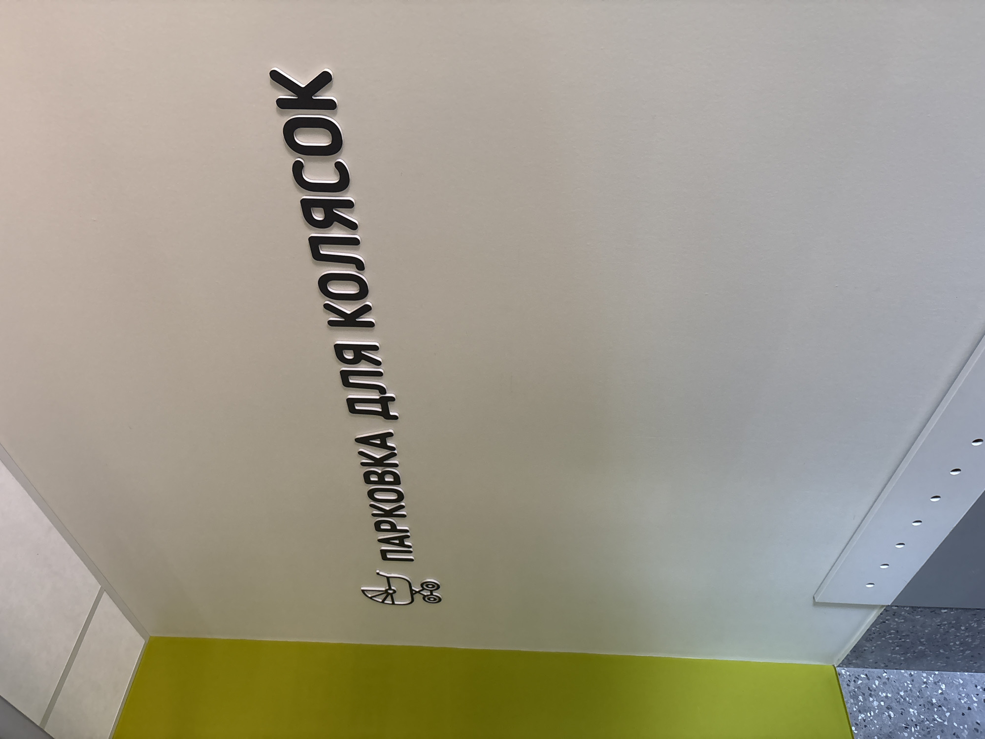 An indoor shot taken from a low angle displays a sign for stroller parking on a wall in Veliky Novgorod, Russia. The primary subject is the vertically oriented text, rendered in black, raised 3D lettering with a subtle white outline or shadow, which reads "ПАРКОВКА ДЛЯ КОЛЯСОК." This translates from Russian as "PARKING FOR STROLLERS." Below the text, a silver-colored pictogram of a baby stroller reinforces the sign's message.
The wall where the sign is mounted is a plain, light off-white or very light grey. A vibrant lime green section forms a base along the bottom of the wall, contrasting with the lighter upper portion. To the right, a grey structural element, possibly a ceiling panel or a decorative strip, features a row of small, circular recessed lights or holes. Further right and towards the bottom, a dark grey floor with a speckled, terrazzo-like texture is visible. The scene appears clean and modern, suggesting a public or commercial space such as a shopping center or a family-friendly facility. No people are present, and the lighting is even, indicating an indoor setting with artificial illumination.