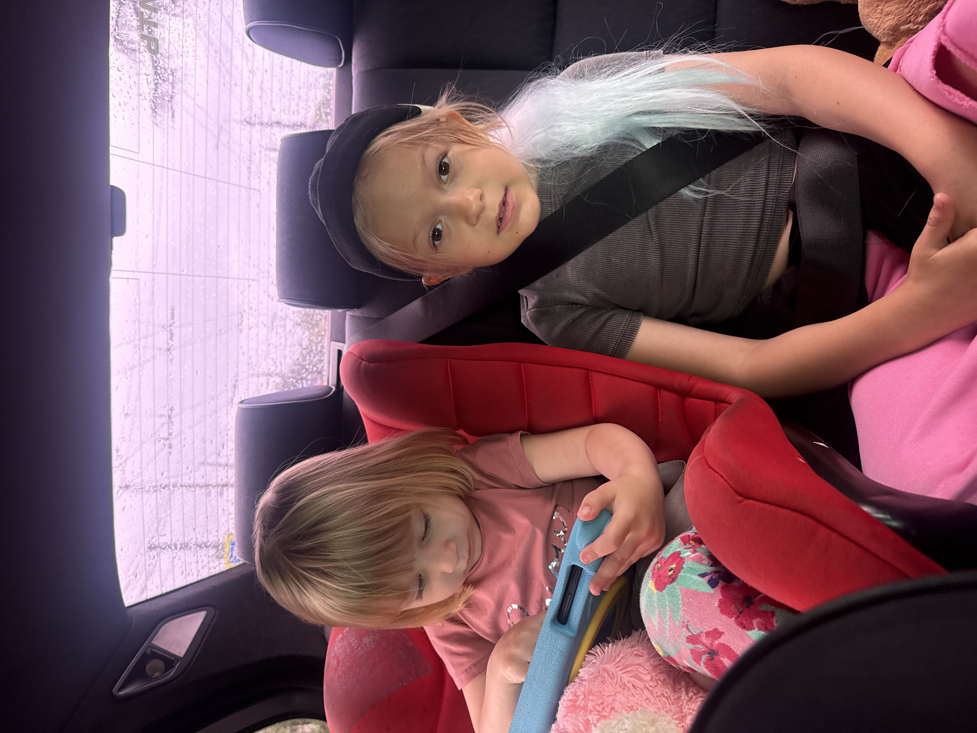 Two young children are inside the back seat of a car. One child, wearing a black cap and a grey shirt, is buckled in with a seatbelt. This child has long blonde hair with strands of blue. The second child, in a pink shirt, sits in a red car seat, looking down and holding a blue electronic device. A floral pillow is visible next to the car seat. The car window behind them shows raindrops on the glass, obscuring the outside view. The interior is mostly dark, with the car seats and the children’s clothing standing out.