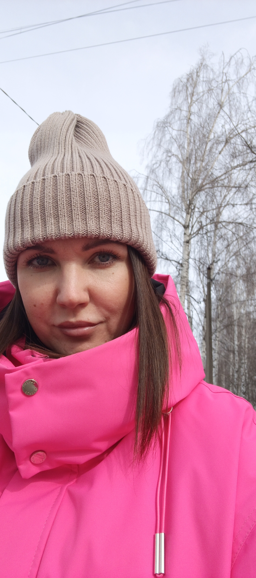 A female individual is depicted in a close-up, selfie-style outdoor portrait in the city of Chaikovsky, Russia. She wears a light beige, ribbed knit beanie hat and a bright neon pink jacket or coat. The jacket features a high collar, possibly part of a hood, secured by a gold-colored snap button on the left side, and has pink drawstrings with silver-colored metallic toggles on the right. The individual has long brown hair, visible beneath the hat and around her shoulders, and light-colored eyes, gazing directly at the camera with a neutral to slightly smiling expression. The background, out of focus, shows an overcast or light gray sky occupying the upper portion. Below the sky, several bare deciduous trees with thin branches are visible, suggesting a colder climate. Thin, dark utility lines traverse the upper left quadrant of the sky.