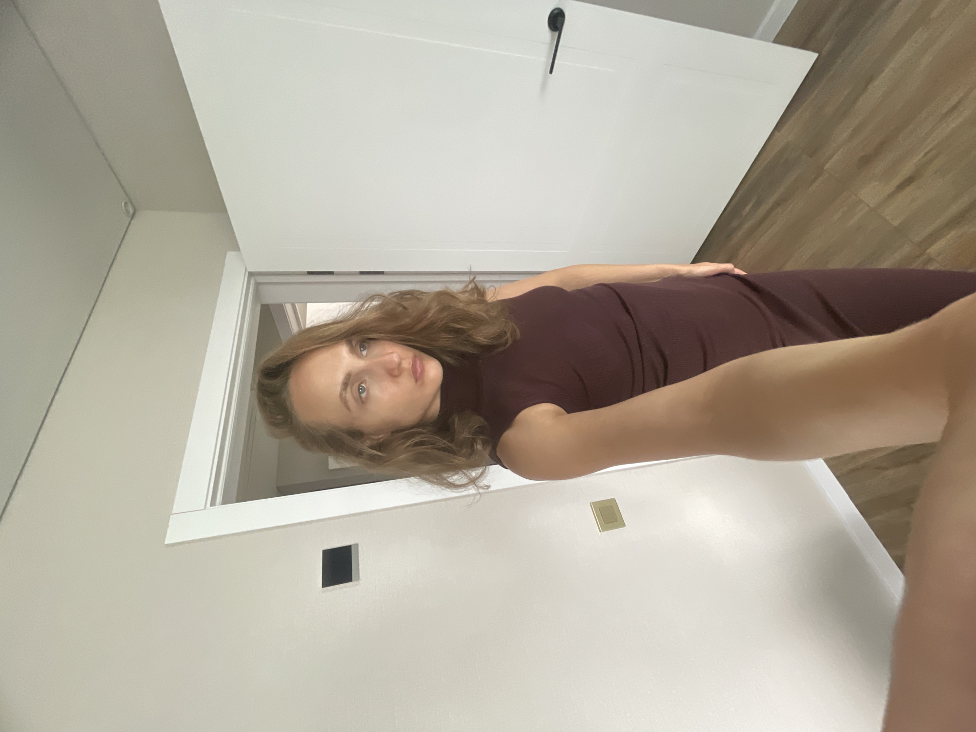 A woman stands in a hallway, extending her arm towards the camera in a selfie pose. She is wearing a sleeveless, dark red, turtleneck dress. Her hair is long and wavy. The hallway features a white door on the right, with a black door handle. There is another doorway visible on the left, leading into another room. The walls are white, and a dark square object is mounted on the left wall. The floor is made of wood-effect laminate.