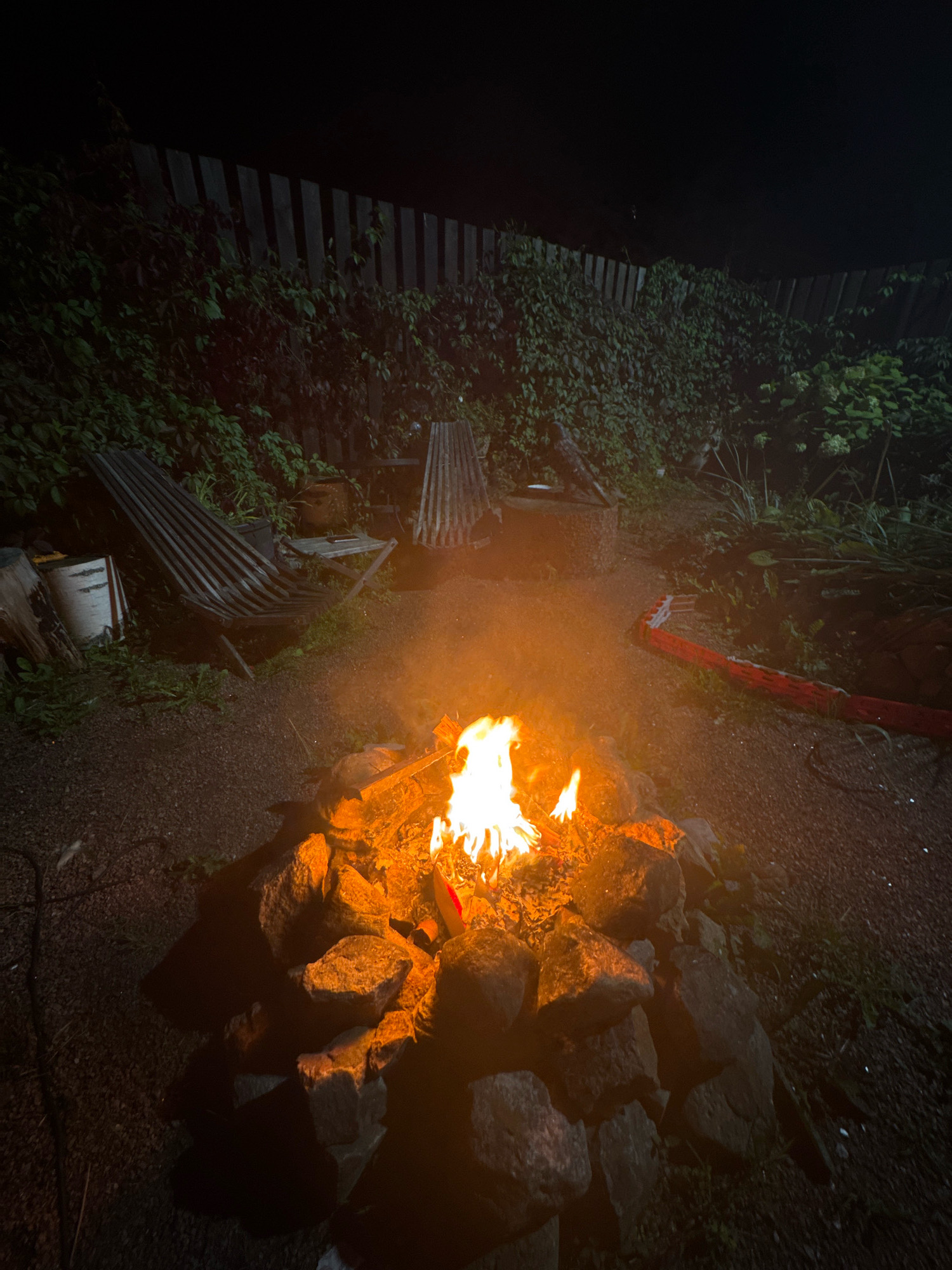 The image depicts a backyard scene at night in Kempelelevo, Russia, featuring a bonfire. The bonfire is centrally located, contained within a ring of large stones. Flames are visible, casting an orange glow on the surrounding rocks and smoke. In the background, a wooden fence lines the property, partially obscured by dense green foliage. Two weathered, slatted lounge chairs are positioned on the left side of the frame, near a stack of firewood and a bucket. A red object, possibly a sleeping bag or mat, lies on the ground to the right of the bonfire. The ground is covered in dark gravel and sparse vegetation. The overall atmosphere is dark, with the fire being the primary light source.