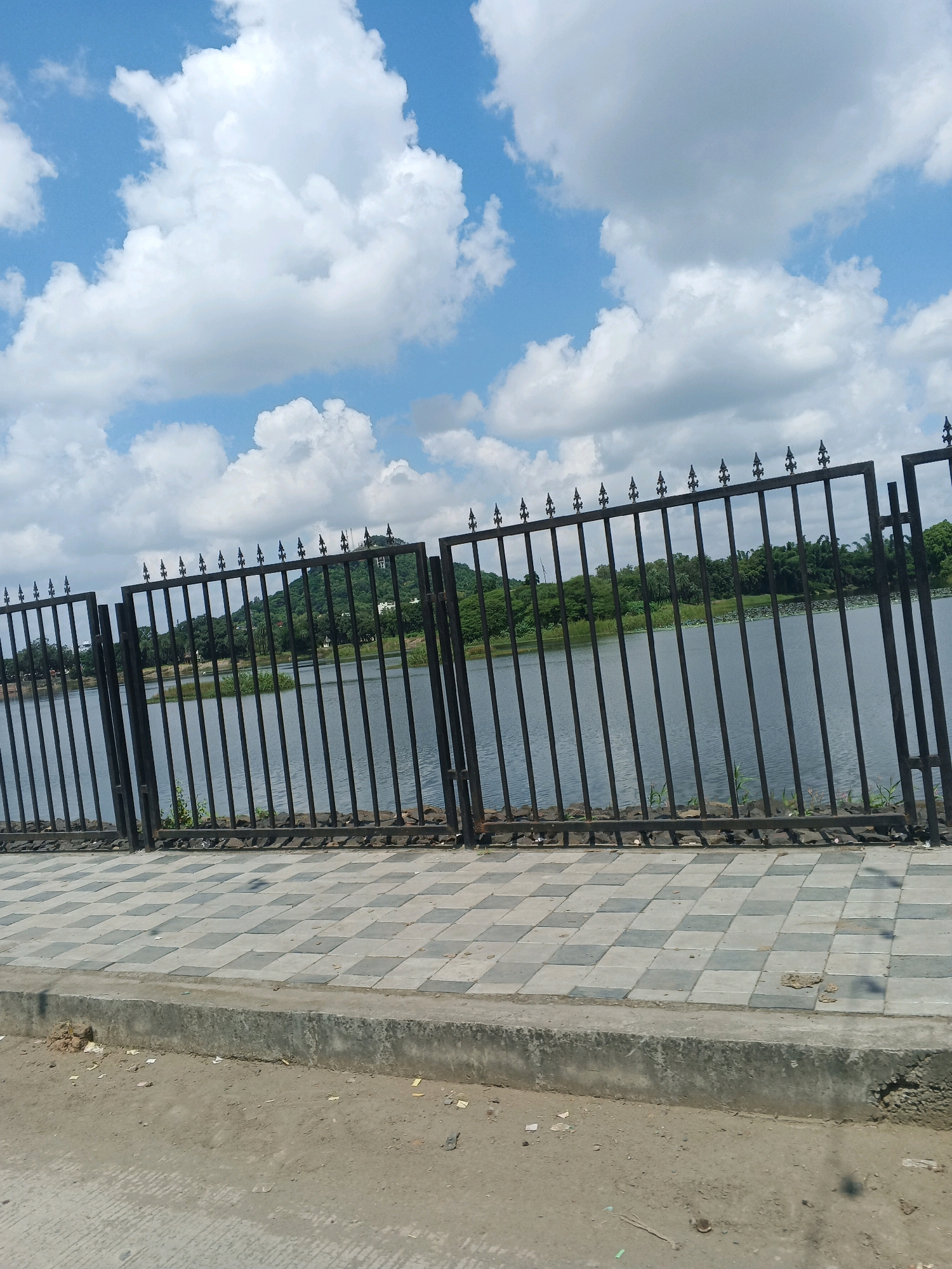The image shows a black metal fence bordering a calm body of water. Beyond the water, a lush green hill is visible, topped with a white structure. The sky above is blue with scattered white clouds. In the foreground, a checkered paved walkway runs alongside the fence, with a concrete curb and a dirt area to its left. The scene is likely a public park or waterfront area in the city of Dewas, India, given the context provided. There are no people or animals visible in the image.