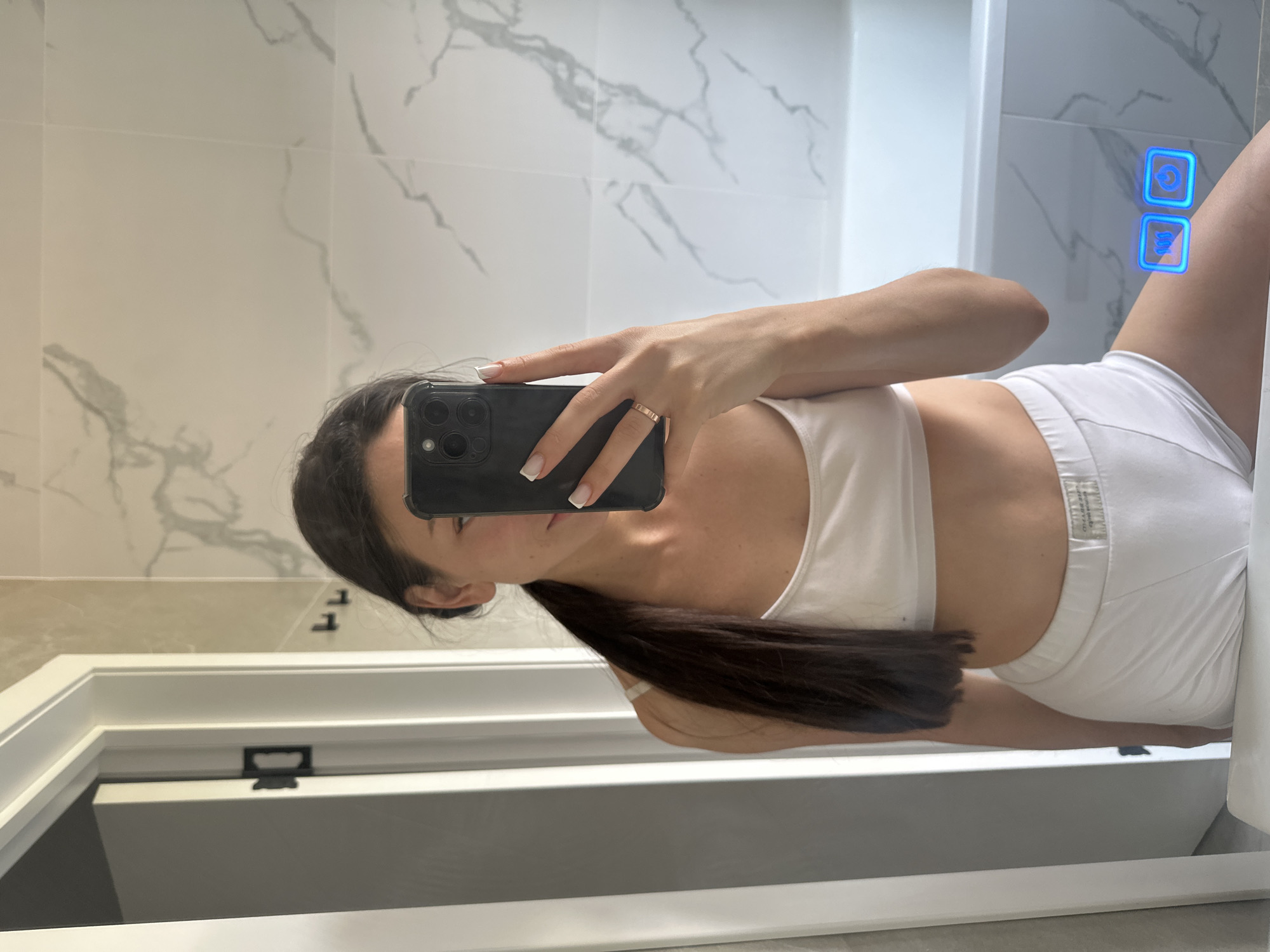 The image is a mirror selfie taken in a bathroom. A young woman with dark hair pulled into a ponytail is wearing a white sports bra and white underwear. She is holding a smartphone in her right hand, which is obscuring part of her face. The bathroom features marble-patterned tiles on the wall and a white bathtub. There are also two illuminated blue buttons on the mirror, one showing "100" and the other a power icon. The location is identified as Estachi, Russia, but this information is not visually present in the image itself.