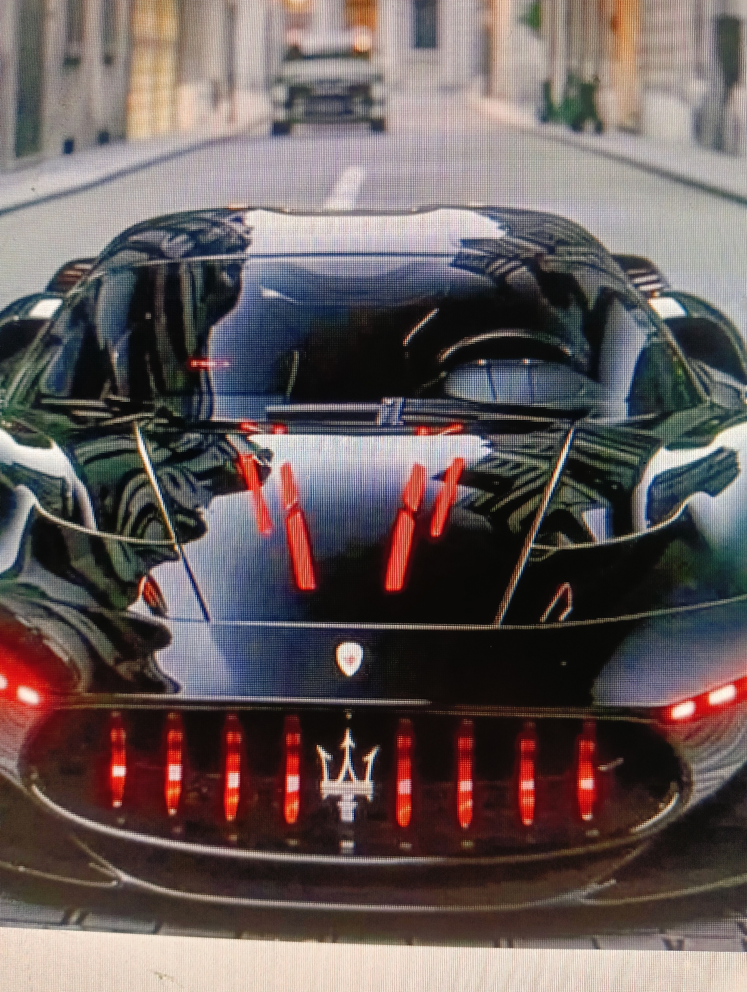 The image shows the front of a black Maserati MC20 car. The car has red vertical accents on the hood and red taillights arranged vertically within a black grille. The Maserati trident logo is visible on the grille. The car is parked on a street that appears to be in a city. The background is out of focus but shows buildings and other vehicles. There are no people visible in the image. The scene is a close-up shot of the car's front.