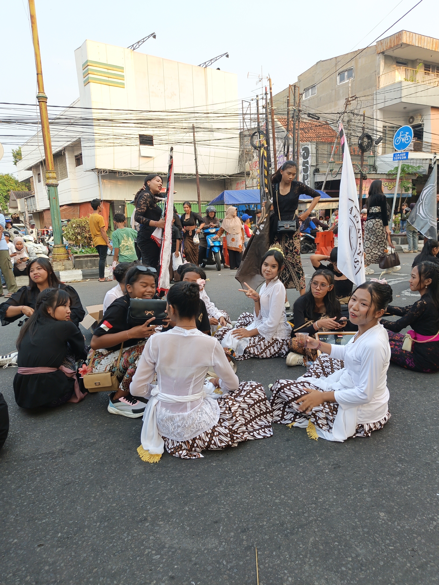 This image depicts a group of young Indonesian women dressed in traditional clothing, sitting on an asphalt street in Klaten, Indonesia. The women, appearing to be of various ages, are adorned in white and black traditional attire, characterized by ornate batik skirts and blouses. Some are wearing white tops with lace detailing and white sashes tied around their waists. Others are in black lace tops and batik skirts. Several women are holding flags, one of which is white with text, and another is black.
The setting appears to be a street in an urban area, with buildings visible in the background. The architecture suggests a commercial or residential district, with a mix of modern and older structures. Power lines and street lamps are also present, indicating a developed area. In the background, other people can be seen, some standing and observing, while others appear to be participating in the event. A blue motorcycle is parked on the right side of the image, and a bicycle lane sign is visible on a pole. The ground is paved with asphalt, and some debris or small objects are scattered on it. The overall atmosphere suggests a cultural event or parade taking place on the street. The lighting indicates it is daytime, likely late afternoon given the angle of the light.