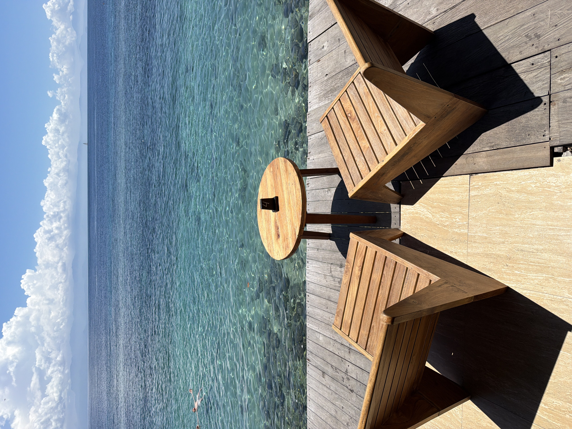 The image depicts a seaside setting, likely in Batununggul, Indonesia, where clear turquoise water meets a wooden deck furnished with two wooden chairs and a small round table. A person is visible in the water. The day is bright with scattered clouds, casting shadows on the deck. The scene suggests a relaxing atmosphere with seating for enjoying the ocean view.