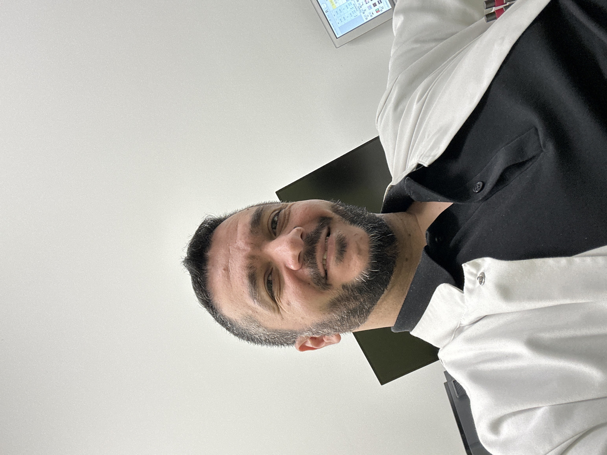 An image, captured from a low, upward angle, features a man looking directly at the camera with a slight smile. The man, who appears to be middle-aged, has short, dark hair with some visible grey at the temples and a full, neatly trimmed beard also showing grey streaks. He is wearing a two-toned shirt, black across the chest and back, with white fabric on the collar and extending over the shoulders and upper arms, possibly a polo shirt or work uniform.
The setting appears to be indoors, likely an office or professional environment, characterized by plain, light-colored walls or ceiling dominating the upper and left side of the frame. In the background, above the man's right shoulder, a monitor or screen is visible. Although small and partially obscured, the screen displays what looks like a digital interface, possibly a spreadsheet or application, with various icons and some faint alphanumeric characters including "0.00" and an "R." The overall impression is one of a candid moment captured within a workspace. The location context provided is Berlin, Germany.