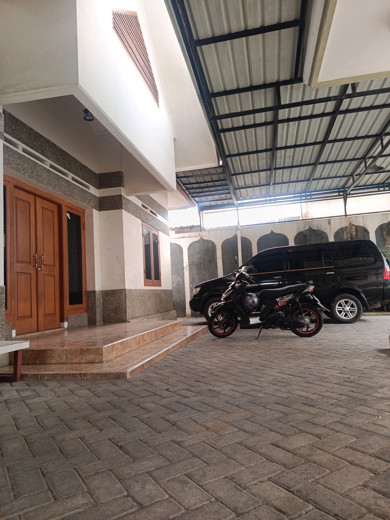 The image depicts a street-level view of a building's entrance and a covered parking area in Kebonagung, Indonesia. The building facade features double wooden doors, a window, and textured stone accents. A tiled staircase leads up to the entrance. To the right, a black van and a black motorcycle with red accents are parked under a metal-framed corrugated metal roof. The ground in front of the building and parking area is paved with interlocking bricks. The environment appears to be a residential or commercial property, with a clean and maintained appearance. There are no people visible in the image.