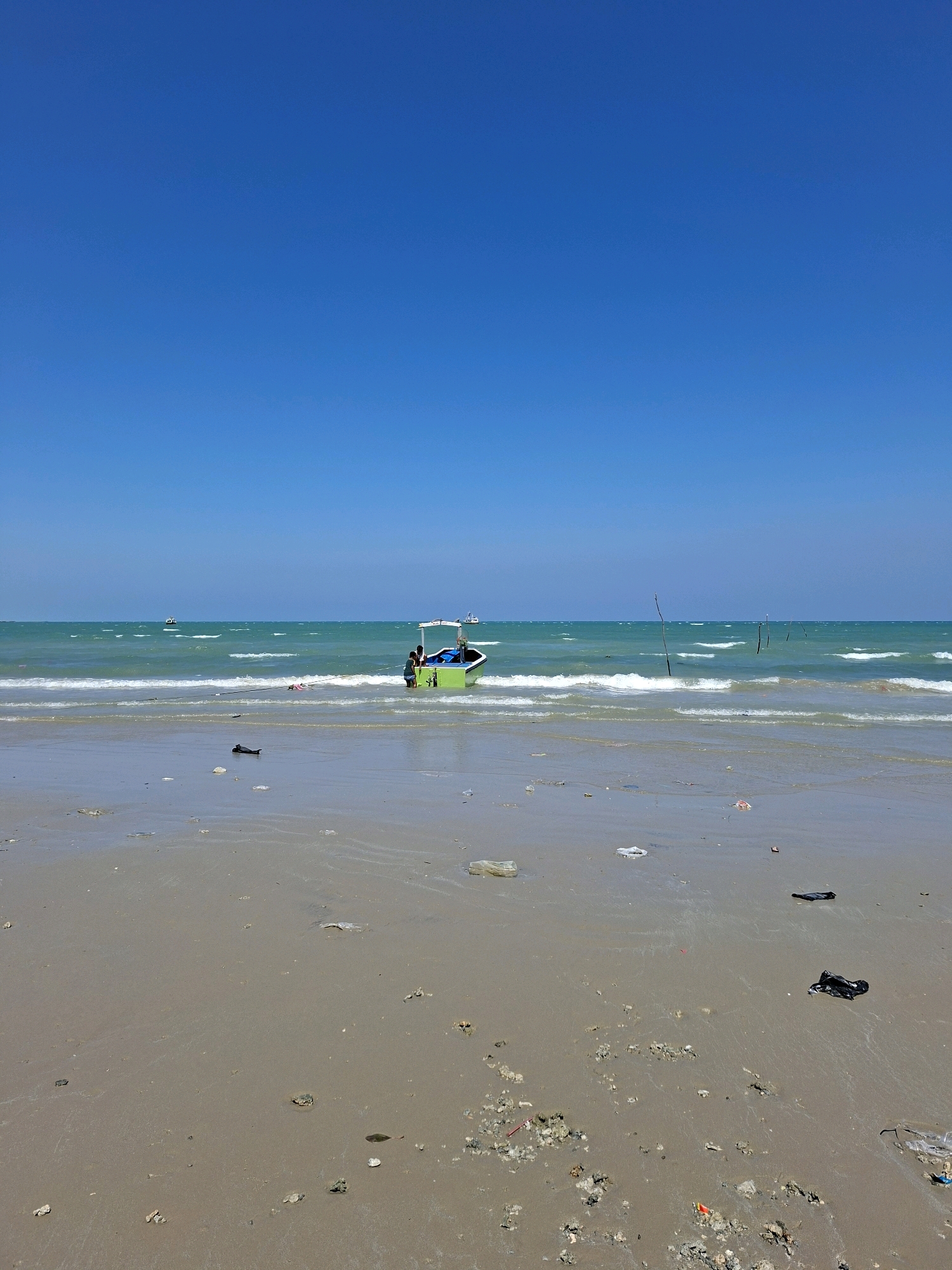 The image depicts a bright, sunny day at a beach in Camplong, Indonesia. The clear blue sky dominates the upper half of the frame. The sea is a vibrant turquoise, with gentle waves breaking on the shore. In the middle ground, a small, lime-green boat with a white canopy is partially submerged in the shallow water. Two individuals, appearing to be children or young adults, are standing in or near the boat. A rope extends from the boat towards the left side of the image. The foreground is a sandy beach littered with small pieces of debris, including plastic bags and what appear to be shells and rocks. In the distance, several other small boats are visible on the horizon. The overall scene is one of a typical beach day, with a touch of environmental concern due to the visible litter on the sand.