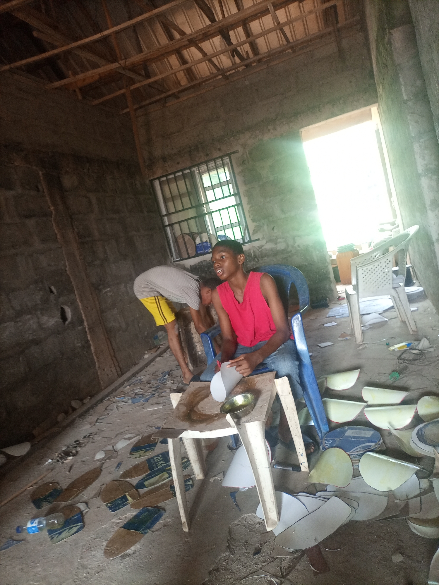 Two young men are in a semi-finished building in Nkpor, Nigeria. The building has a rough concrete block wall and a wooden rafter ceiling. Natural light streams in from an open doorway on the right and a window with metal bars on the left. Scattered on the concrete floor are various shaped pieces of what appear to be ceramic tiles or similar materials, some with blue and brown patterns and text.
One young man, wearing a red sleeveless shirt and blue jeans, sits on a blue plastic chair. He is holding a piece of white material and a small metal bowl. The other young man, wearing a grey t-shirt and yellow shorts, is bent over, examining something on the floor.
On a small white table in front of the seated young man, there is a metal bowl and some white material. To the right of the image, there are white plastic chairs and other objects. The overall scene suggests a workshop or construction site where tile work is being done. The lighting indicates it is daytime, with bright sunlight indicating a clear day. The presence of unfinished construction and scattered materials gives the setting a utilitarian and slightly chaotic feel. No specific emotions are discernible, but the individuals appear focused on their tasks.