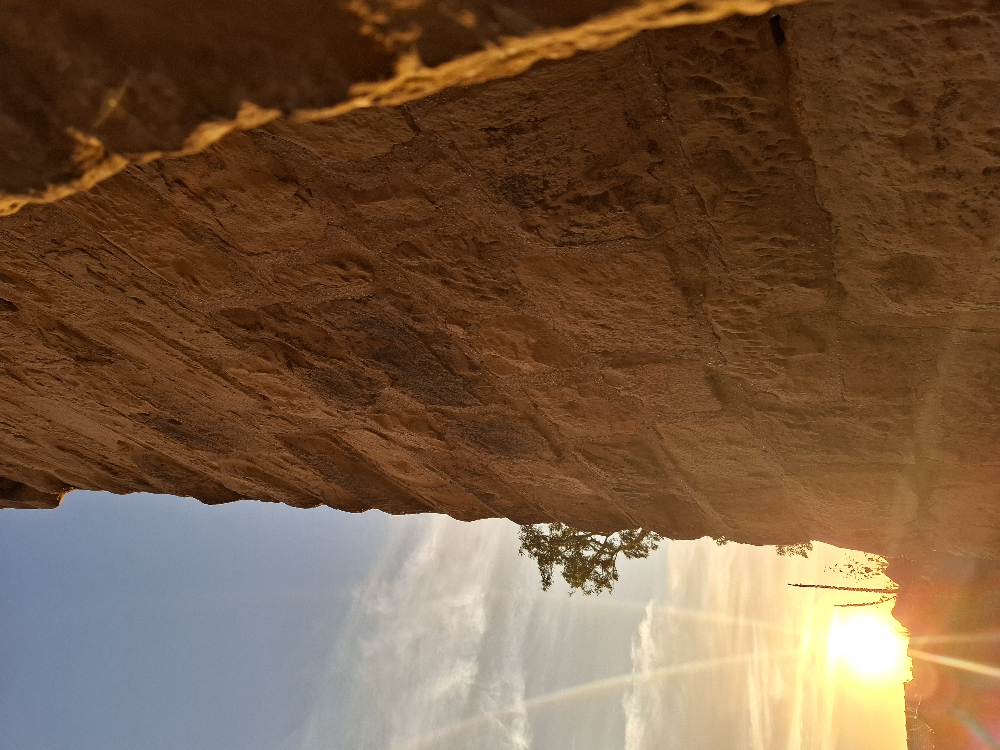 The image captures a low-angle view of a sunlit, rough-hewn stone wall, likely part of a historical structure. The wall dominates the right side of the frame, with its textured surface illuminated by the warm glow of the setting sun. To the left of the wall, the sky transitions from a clear blue at the top to a bright, hazy orange near the horizon, indicating sunset. Sun rays emanate strongly from the left, creating lens flares and a luminous effect. A solitary tree with green foliage is visible in the middle ground, silhouetted against the sky and the wall. In the far distance, faint outlines of buildings suggest an urban or semi-urban environment. The overall atmosphere is one of natural beauty and a sense of time, with the ancient-looking stonework juxtaposed against the dynamic sky. There are no people or animals present in the image. The location is identified as Sinsheim, Germany, suggesting this is a scene from that region.