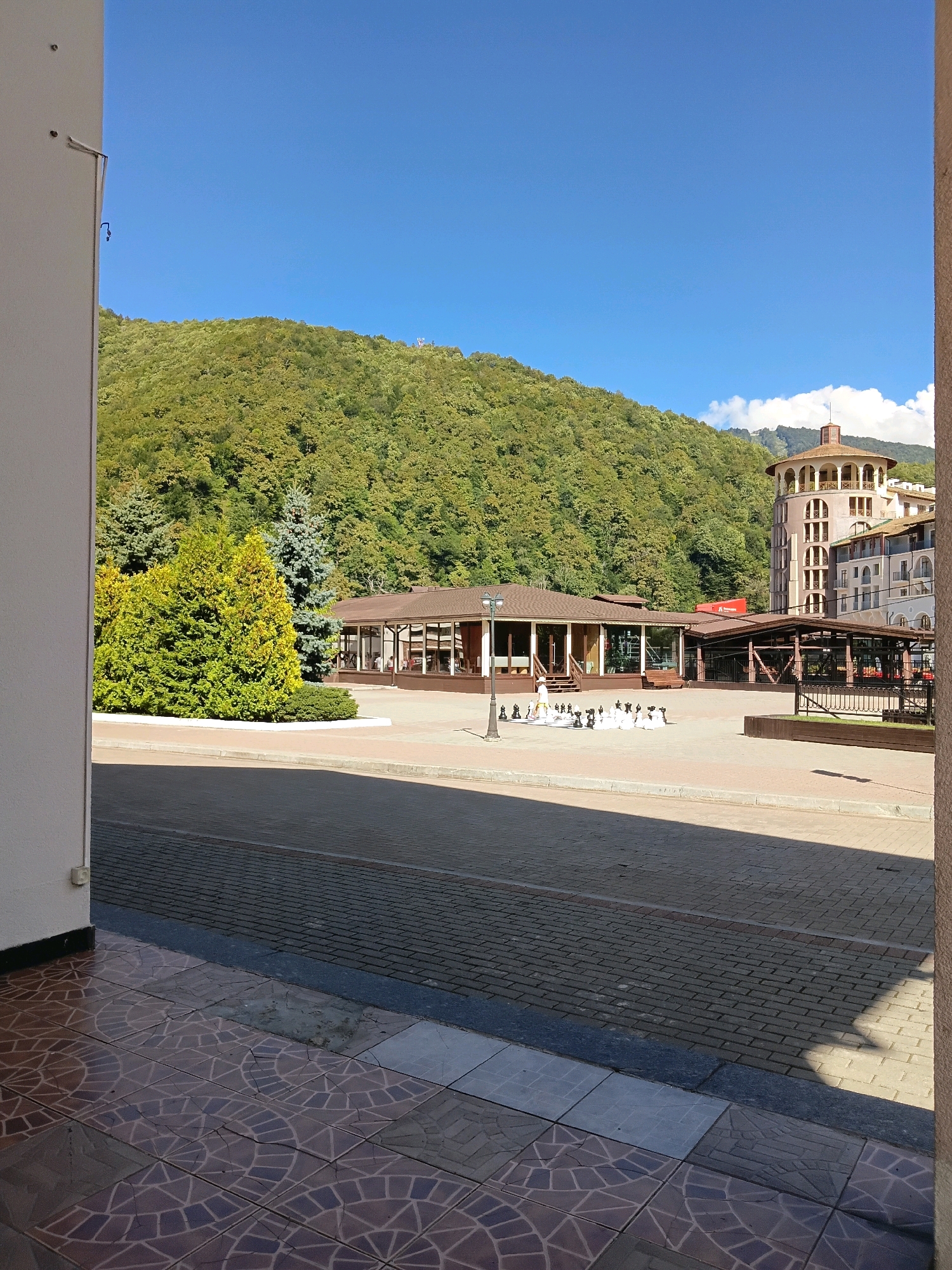 The image captures a sunny day in Estosadok, Russia, featuring a large outdoor chess set set up in a paved plaza. In the background, a lush, green forested mountain rises under a clear blue sky. To the right of the mountain, a multi-story building with a distinctive circular tower is visible. In the foreground, a tiled patio leads to a paved area where the chess pieces are arranged. A single person, dressed in white, appears to be interacting with the chess pieces. Several other buildings, including a single-story structure with large windows, are also present in the scene. The overall atmosphere suggests a recreational or tourist area.