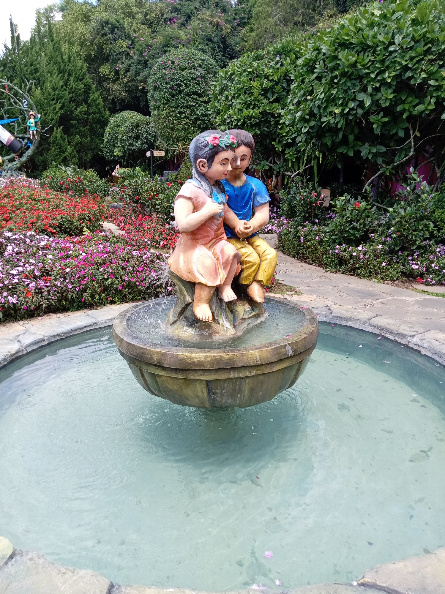In Da Lat, Vietnam, a stone fountain features a statue of a young boy and girl. They are depicted sitting on a rock, facing each other, with their heads close together. The girl wears a pink dress and a flower crown, while the boy is in a blue shirt and yellow pants, also adorned with a flower crown. Their hands are intertwined, and they appear to be holding a small blue bird. The fountain's basin is filled with clear, turquoise water, with ripples indicating a gentle flow. Surrounding the fountain is a vibrant garden filled with a variety of flowers in shades of red, pink, and white, as well as lush green trees and bushes, creating a picturesque and serene atmosphere.